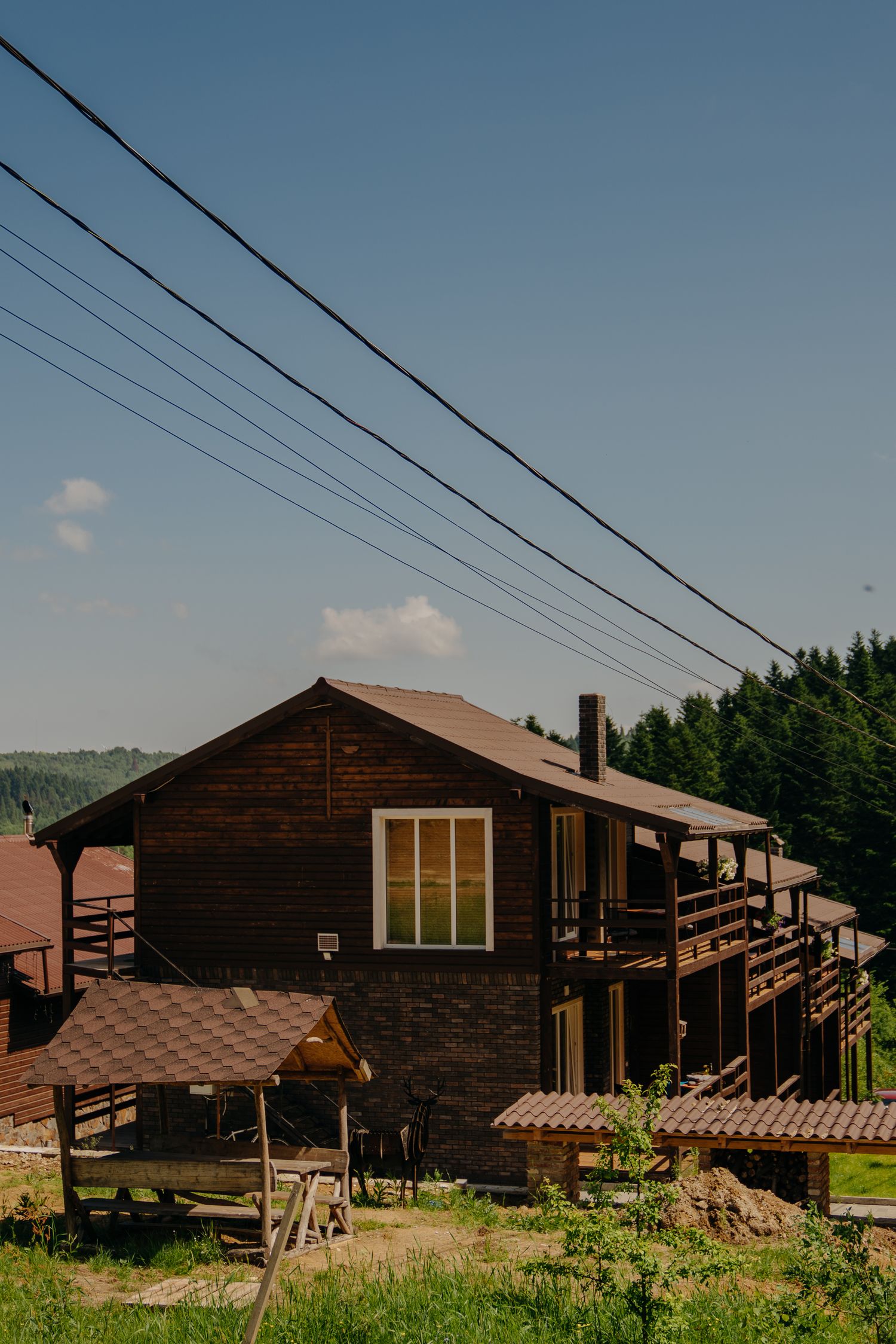 Rustic two-story wooden cabin with balcony surrounded by green trees and overhead power lines in summer.