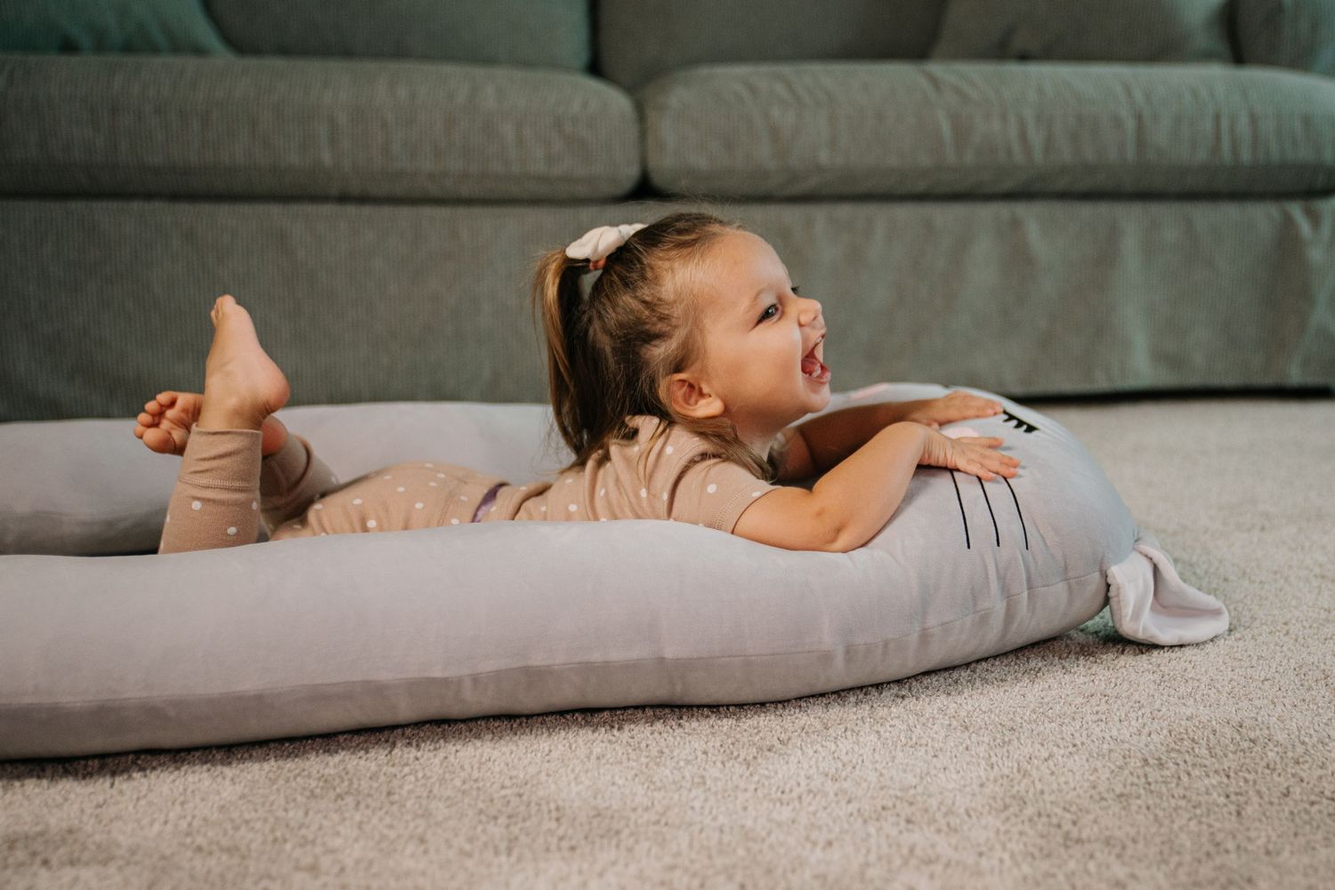 Toddler girl laughing while lying on a large gray body pillow on a carpeted floor near a green sofa.