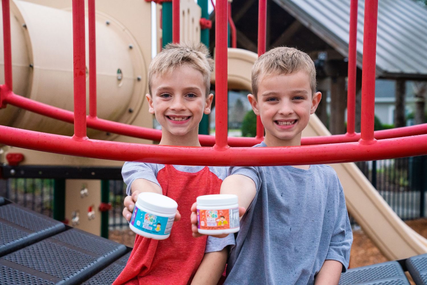 Two smiling boys holding colorful cups on a red playground climbing structure outdoors.