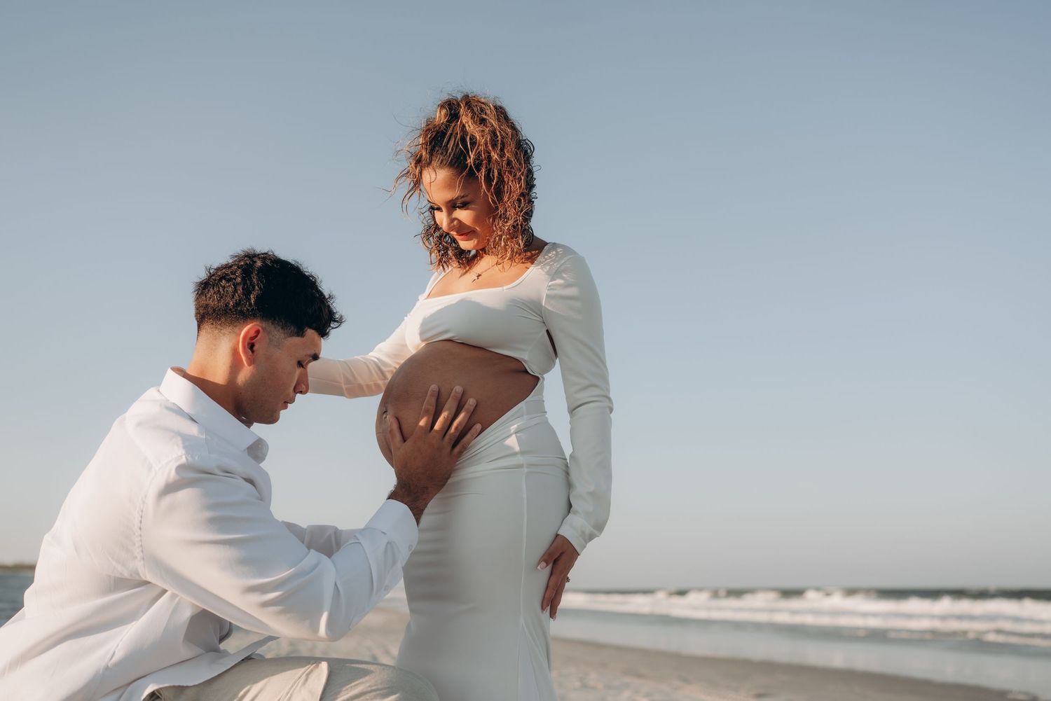 A couple in white outfits embraces on a sunny beach during a maternity photoshoot.