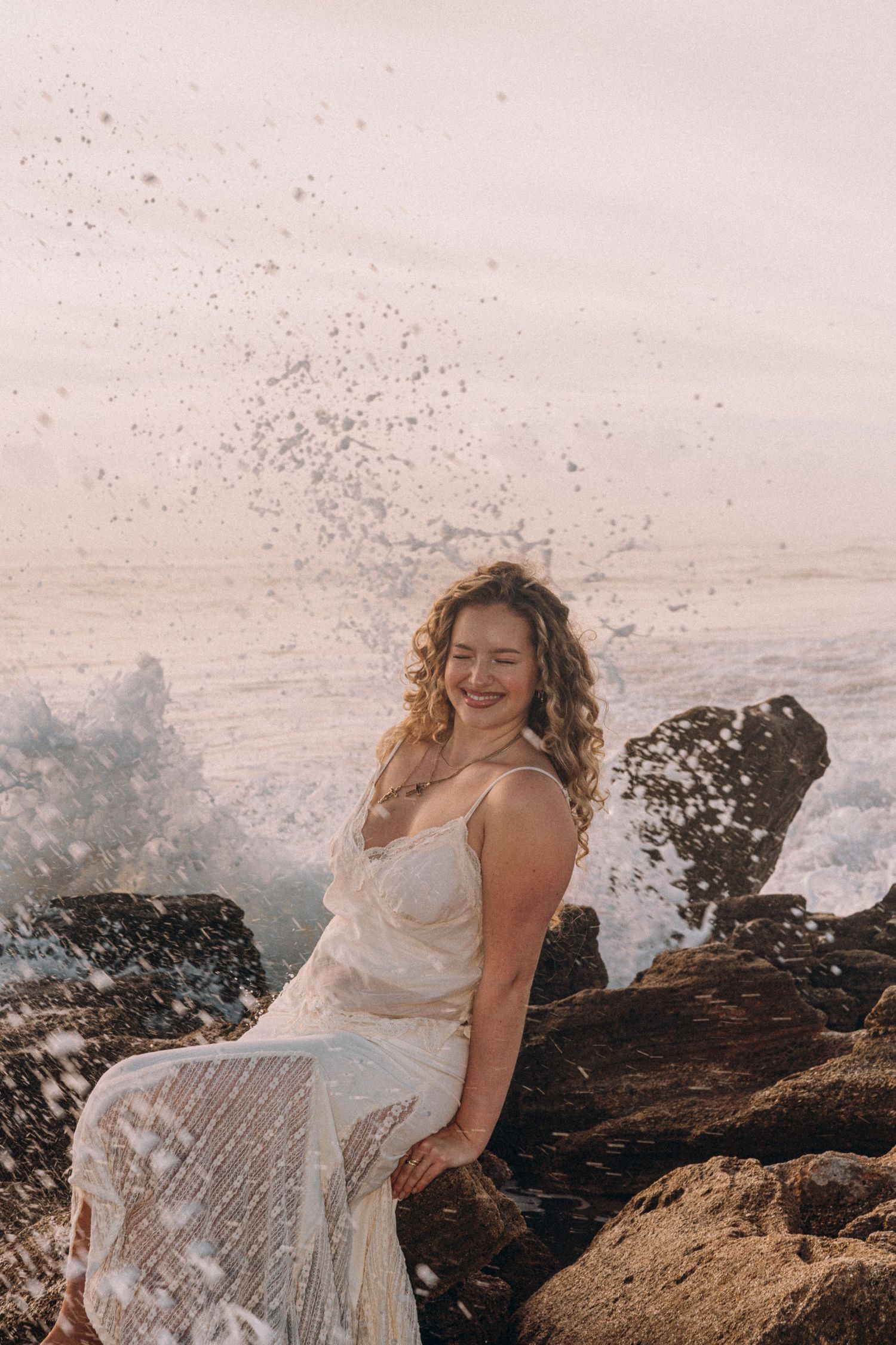 A joyful woman in a white lace dress sits on rocks as ocean waves crash behind her in golden light.
