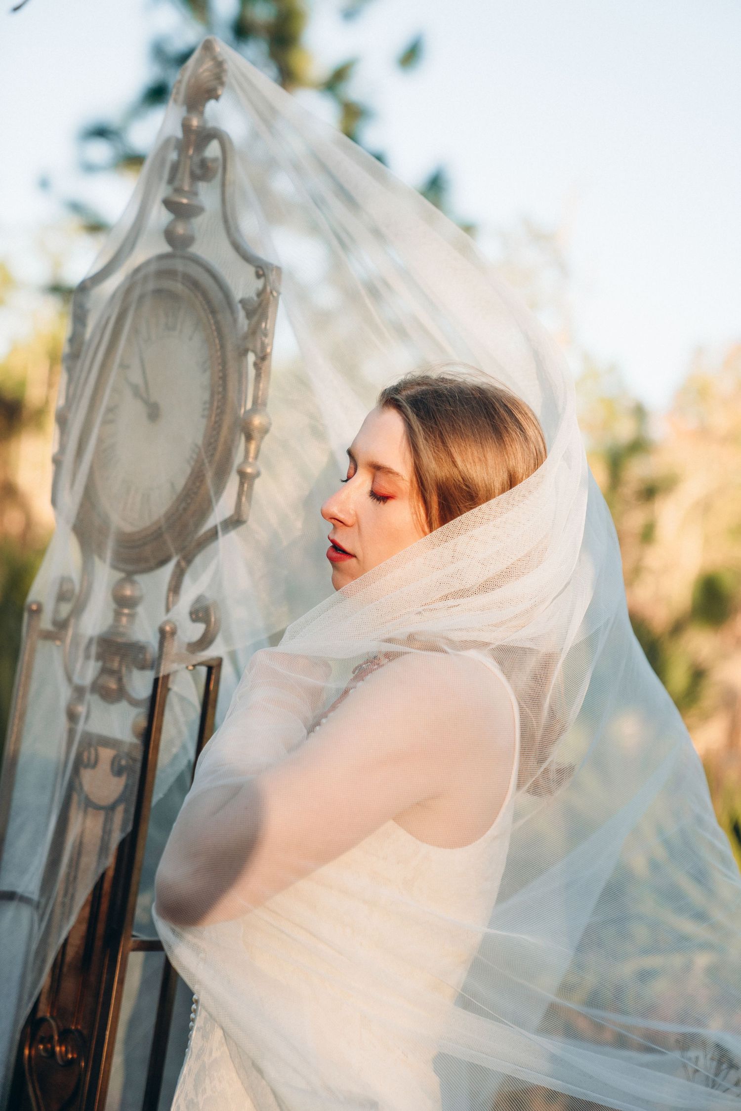 Bride in white wedding dress covered by flowing veil poses outdoors near vintage ornate clock at golden hour.