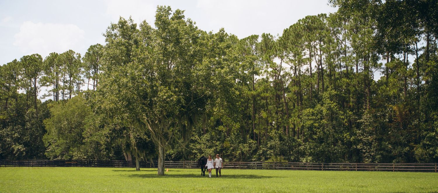 A couple enjoys a peaceful moment in a vast green field with tall trees in the background.