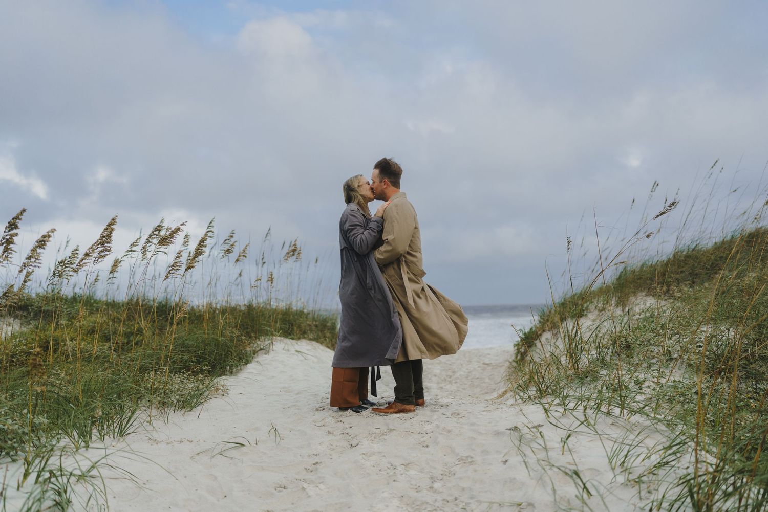 A romantic couple embraces on a sandy beach path lined with sea oats on a cloudy day.