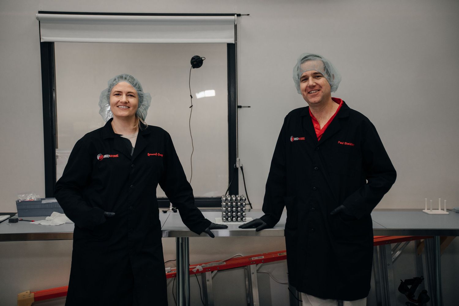 Two professionals in black lab coats and hair nets stand smiling in a clean laboratory environment.