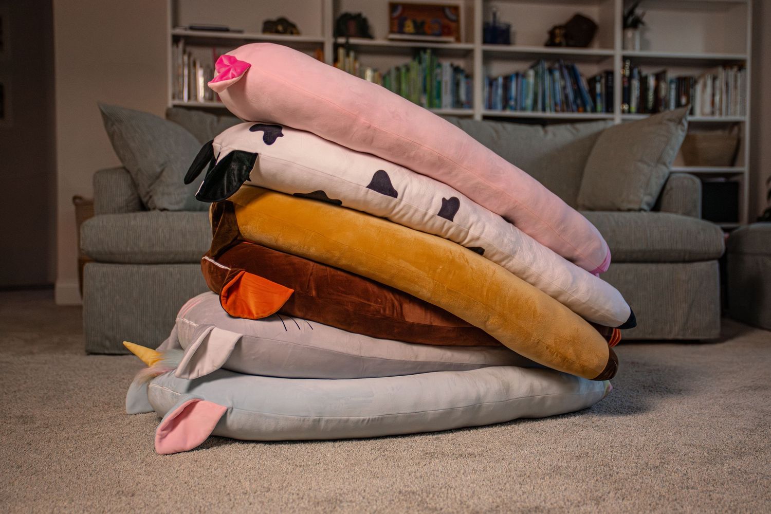 Stack of various large stuffed animal body pillows piled on living room floor near a couch.