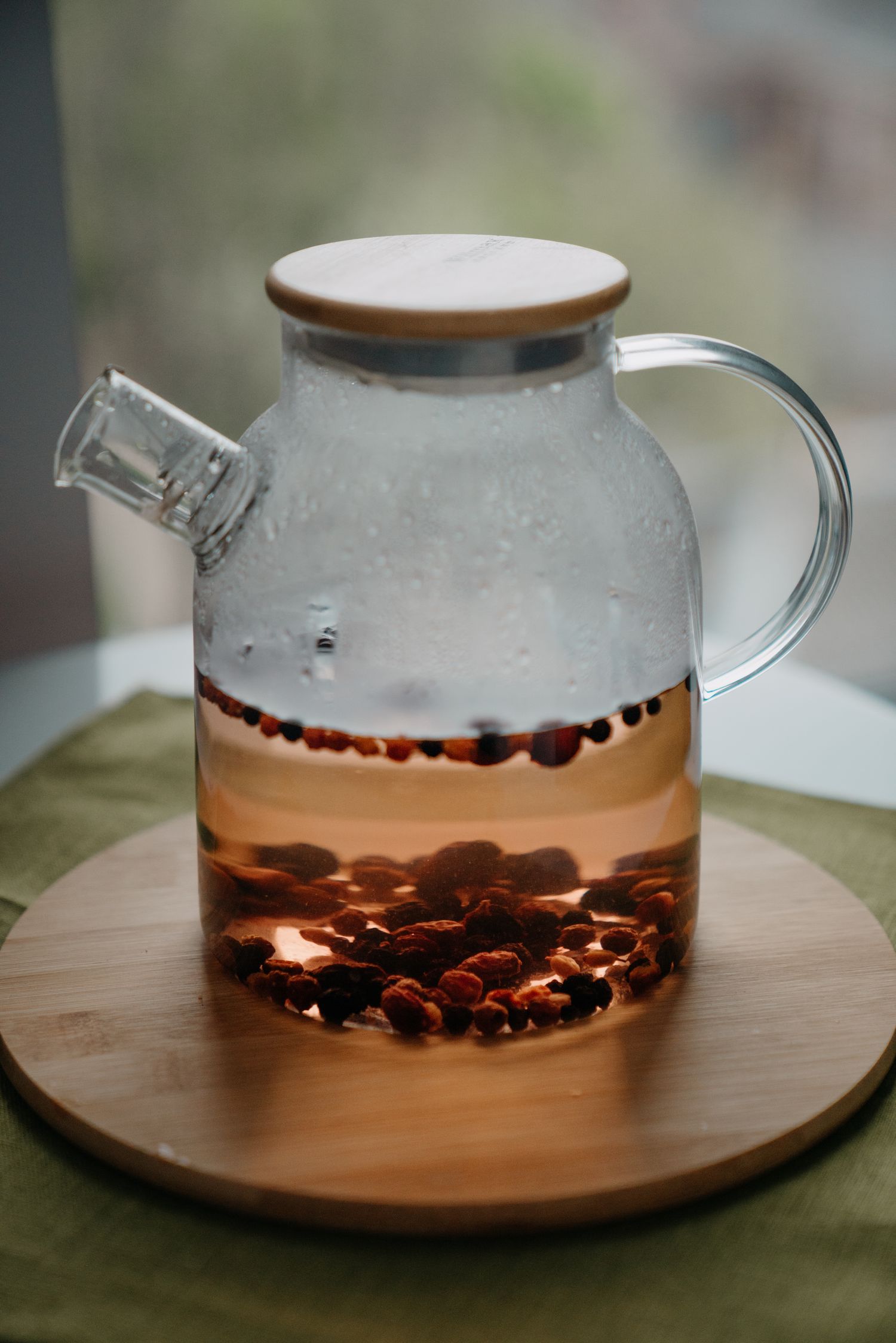 Glass teapot with rose hip herbal tea brewing on a wooden board near a window.