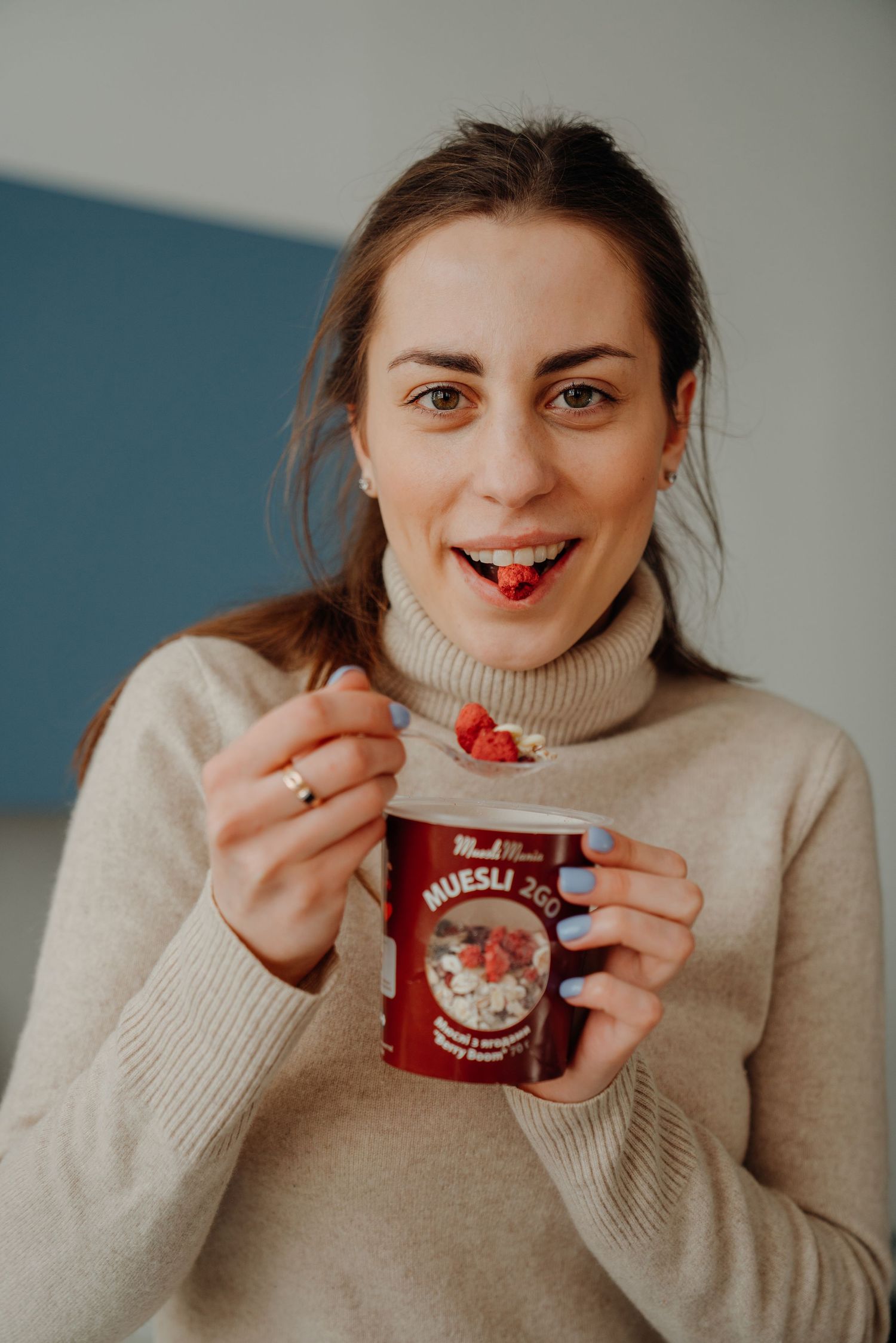 Smiling woman eating berry muesli from a red MuesliMania cup with a spoon.