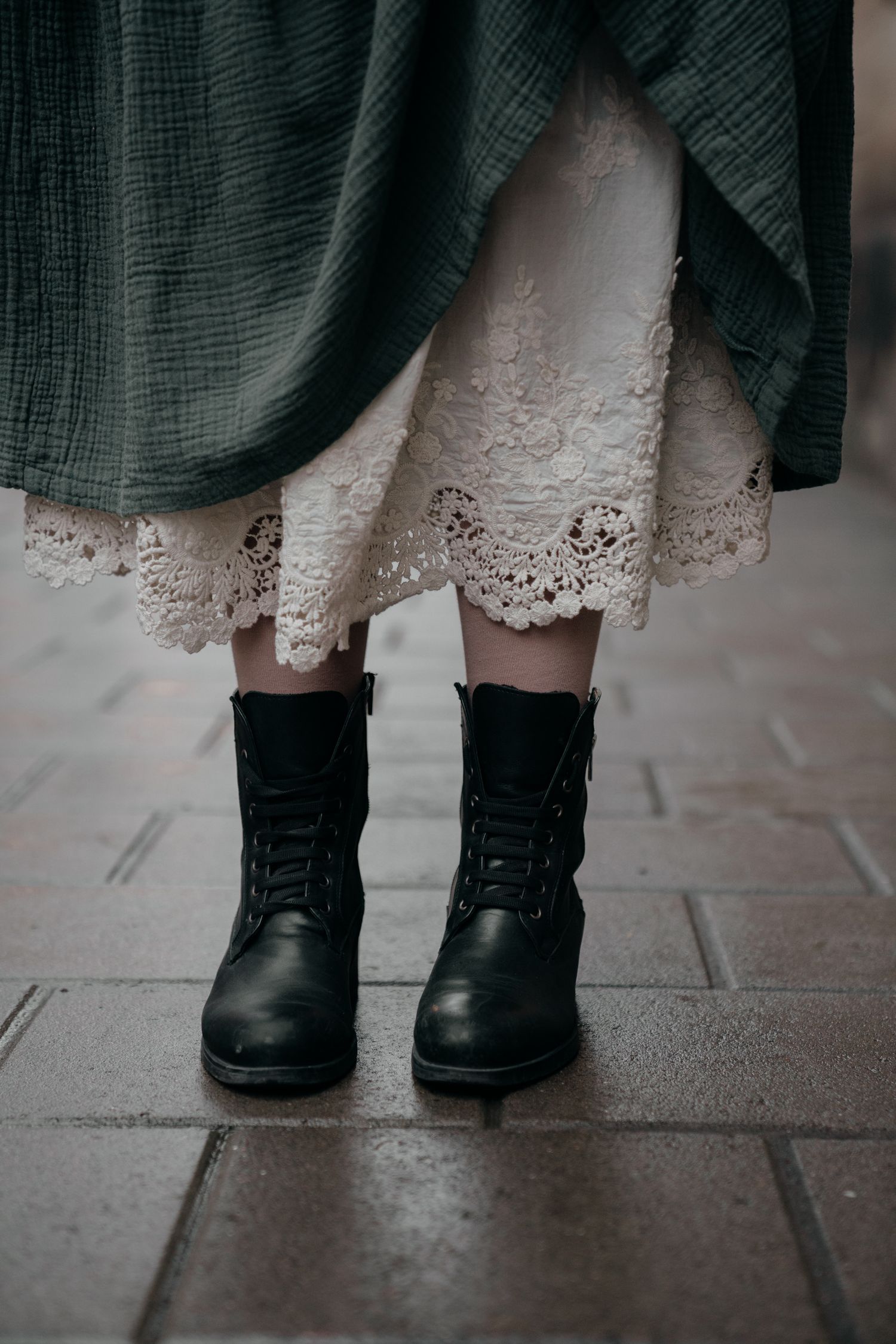 Woman wearing black lace-up boots with a cream lace underskirt and green plaid dress standing on cobblestone.