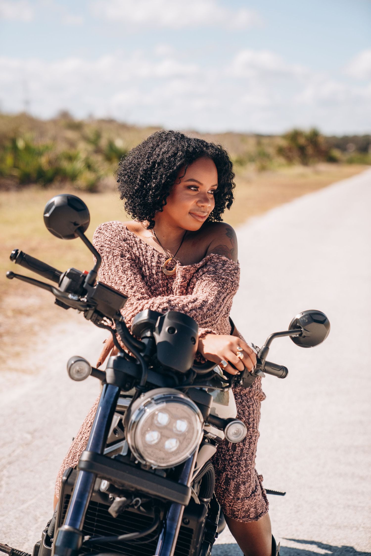 A person posing with a custom black motorcycle on a bright sunny road.
