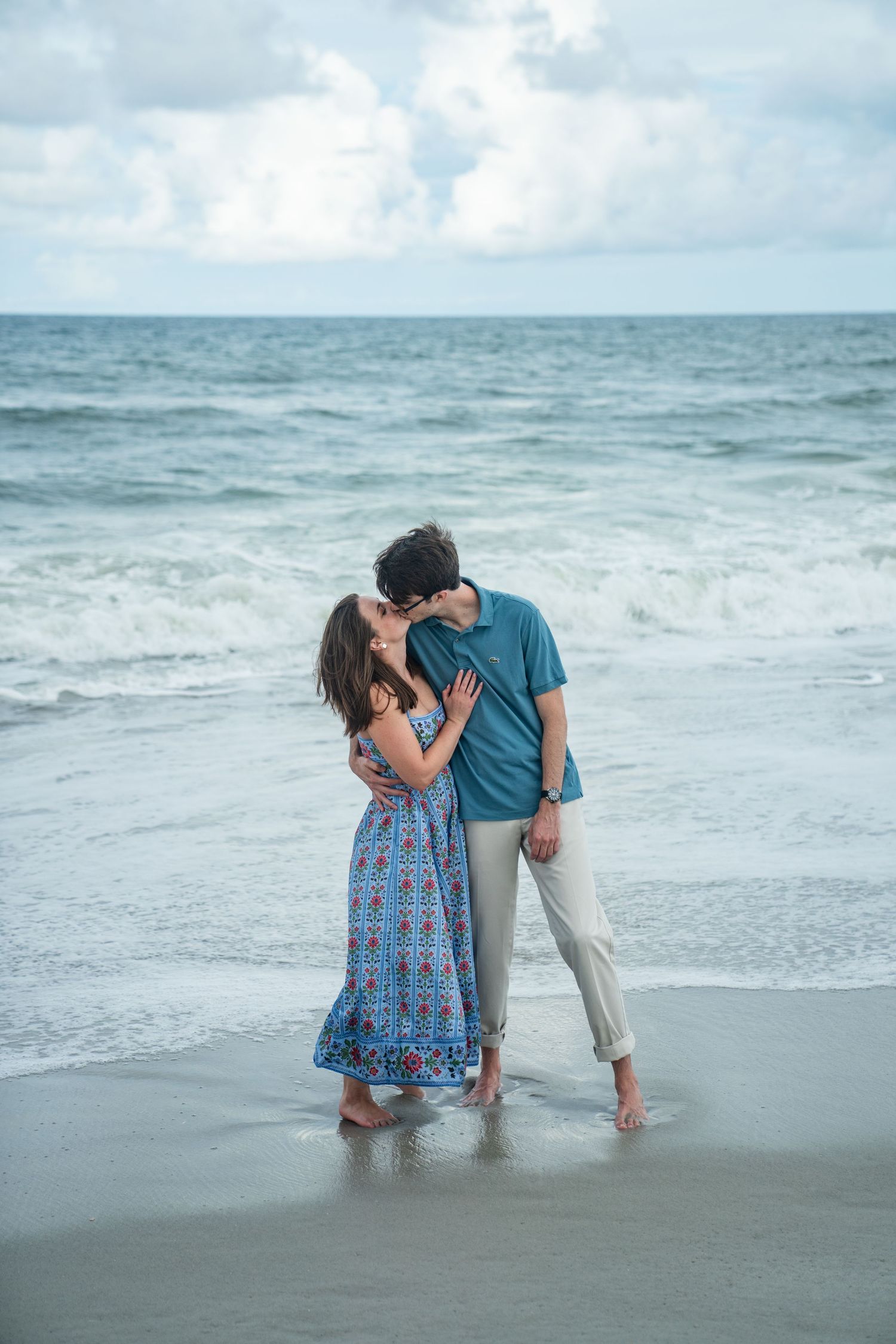 A playful couple dips and dances together on the beach shore with rolling waves and cloudy skies behind them.