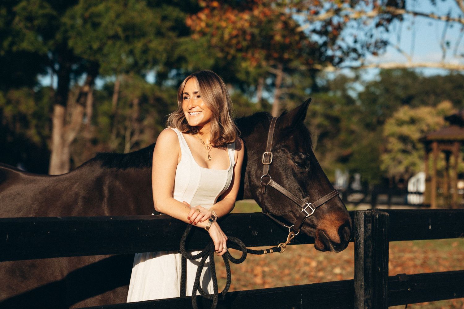 A white-dressed companion stands with a dark horse by a wooden fence at sunset.