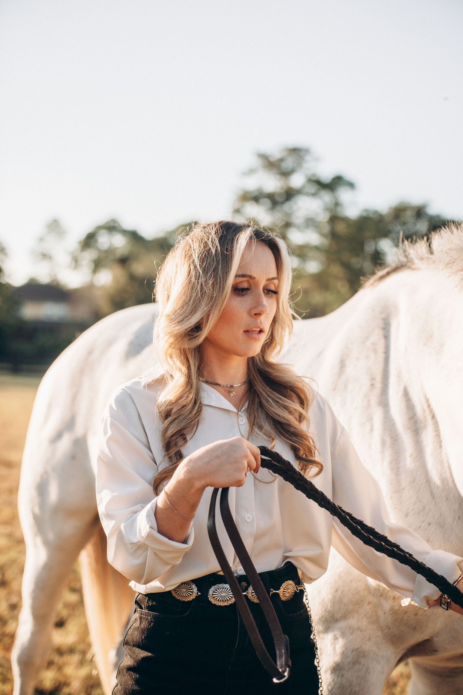 A rider in western attire holds a leather lead rope while standing beside a white horse in a sunlit field.
