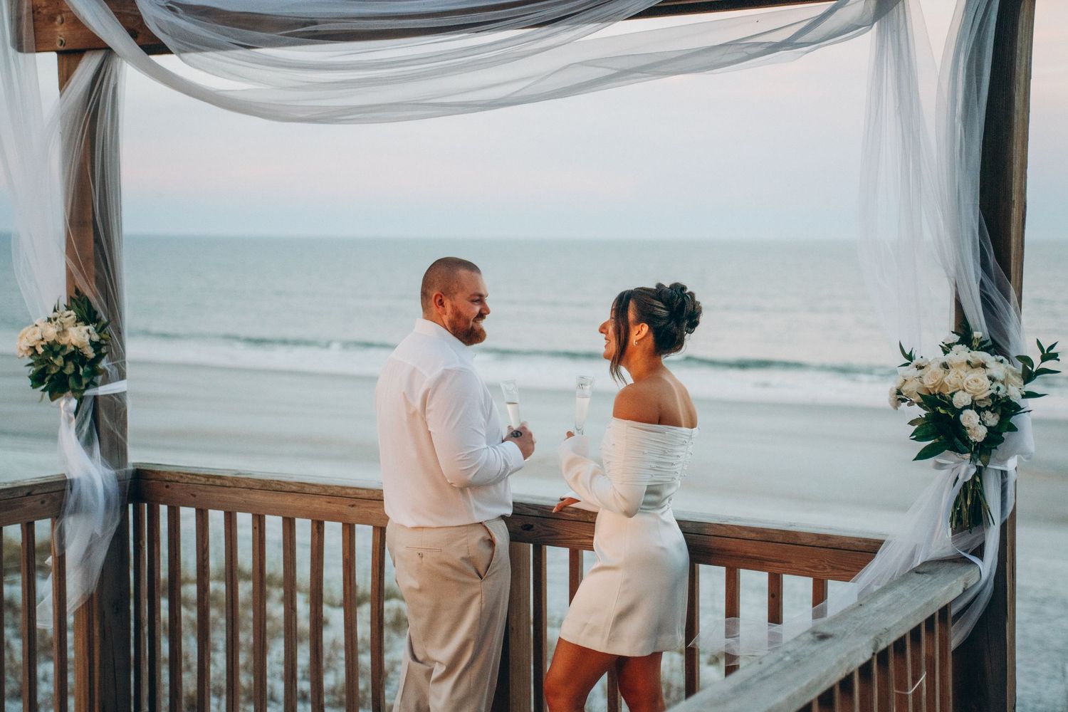 Couple in white attire exchanging vows at a beachside wedding ceremony under a draped canopy at sunset.