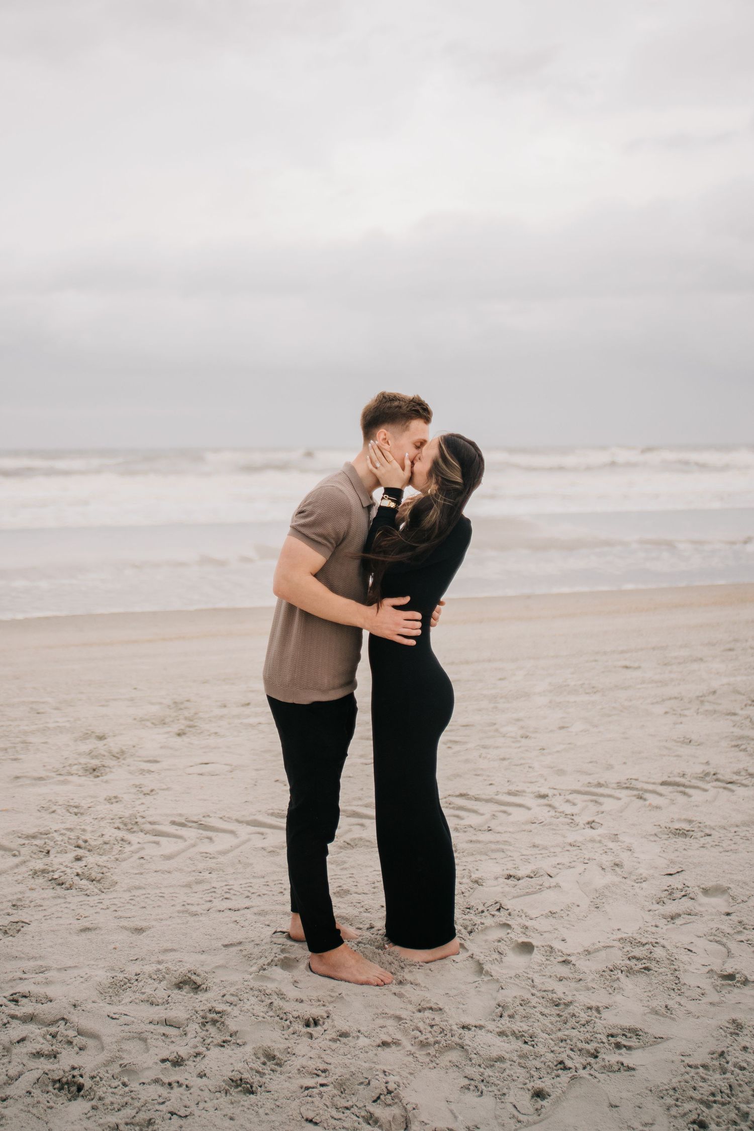 Two people share a loving embrace on a cloudy beach with waves in the background.