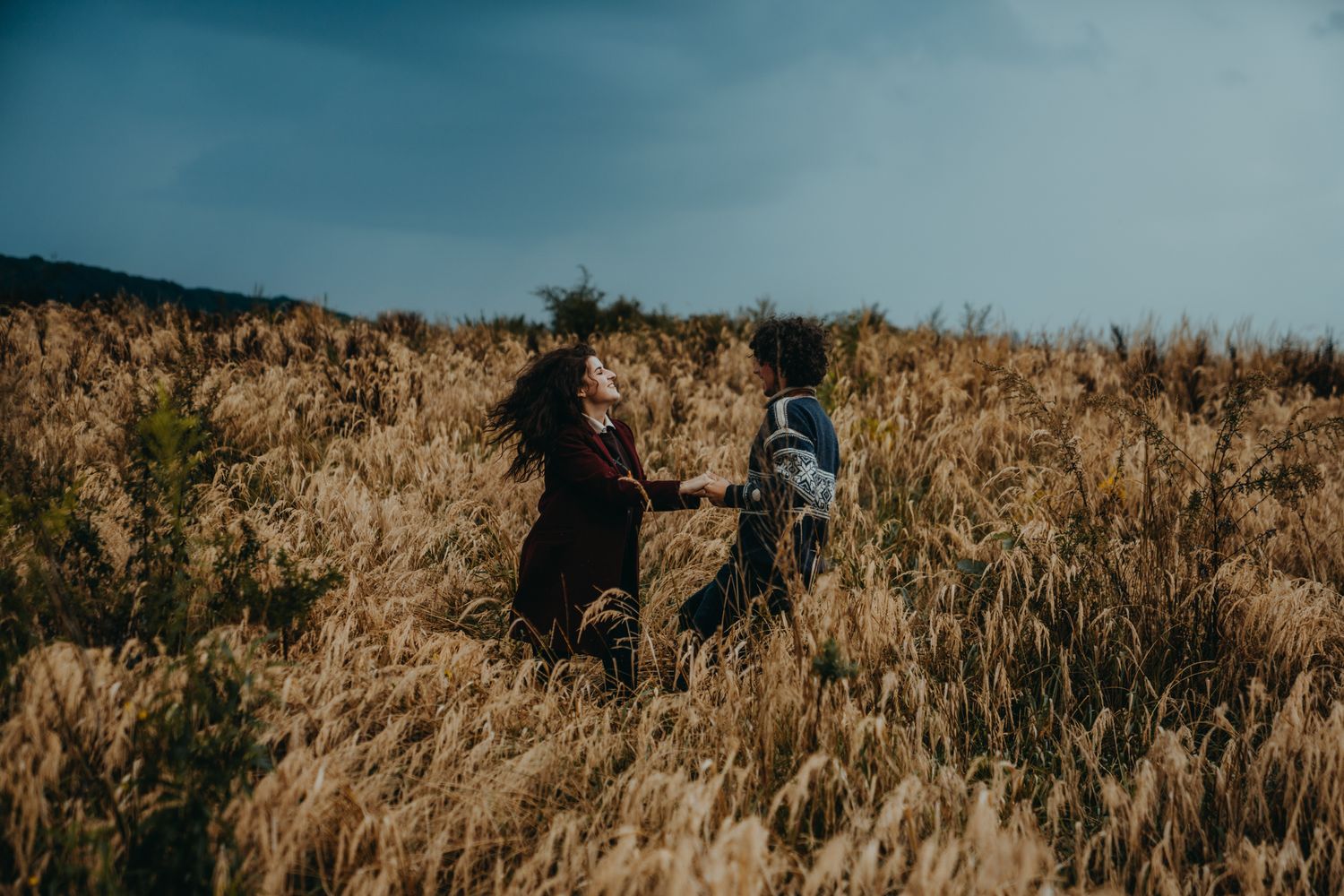 Two figures running through tall grass field against moody sky.