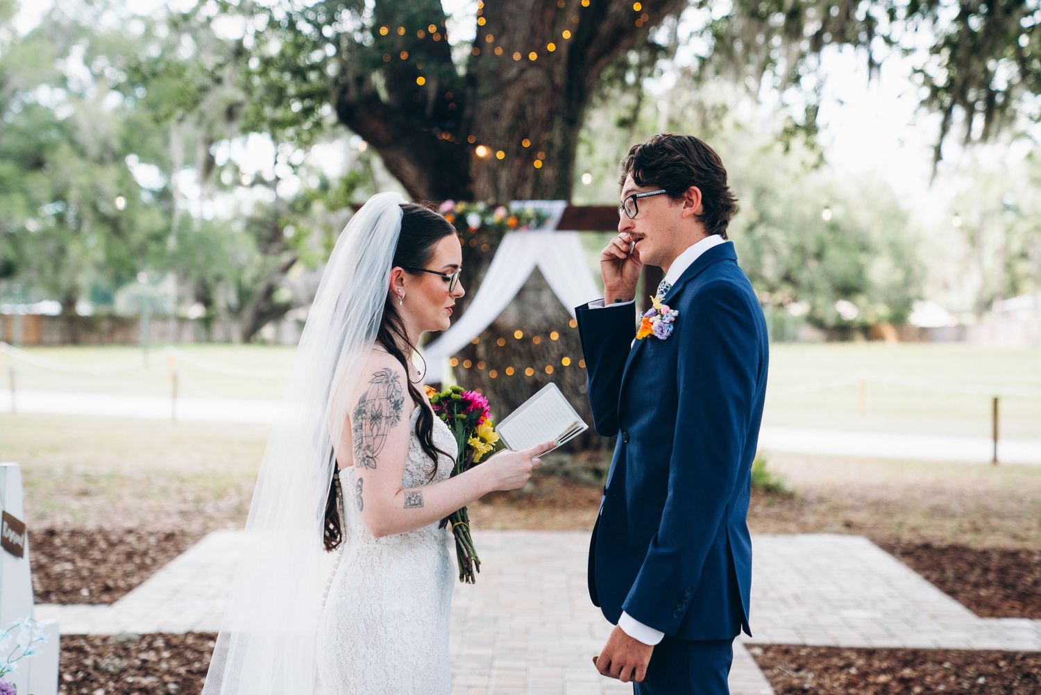 Bride reads vows to groom during outdoor wedding ceremony under oak tree with fairy lights and floral arch.