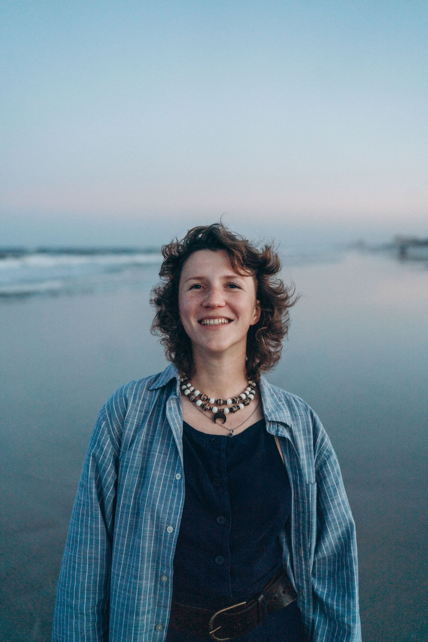 Smiling young woman with curly hair wearing a blue shirt and beaded necklace standing on a beach at dusk.