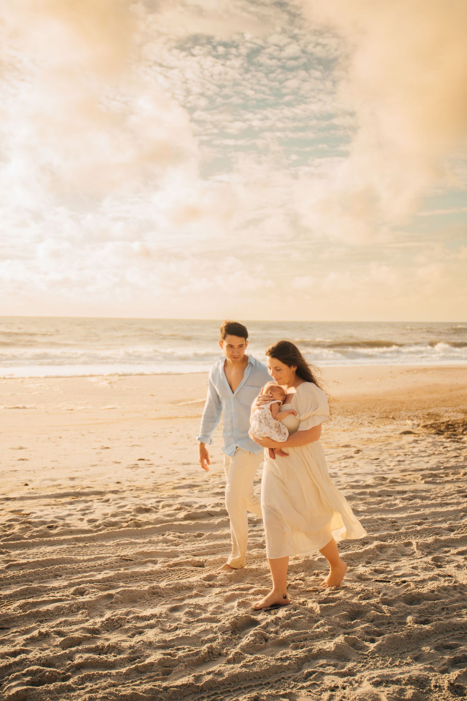 A couple walks on a sunny beach at golden hour, the woman in a white dress cradling a newborn baby.