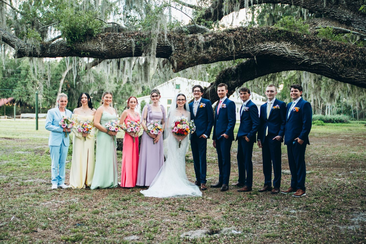Wedding party poses under a large oak tree with Spanish moss, bridesmaids in colorful gowns and groomsmen in navy suits.