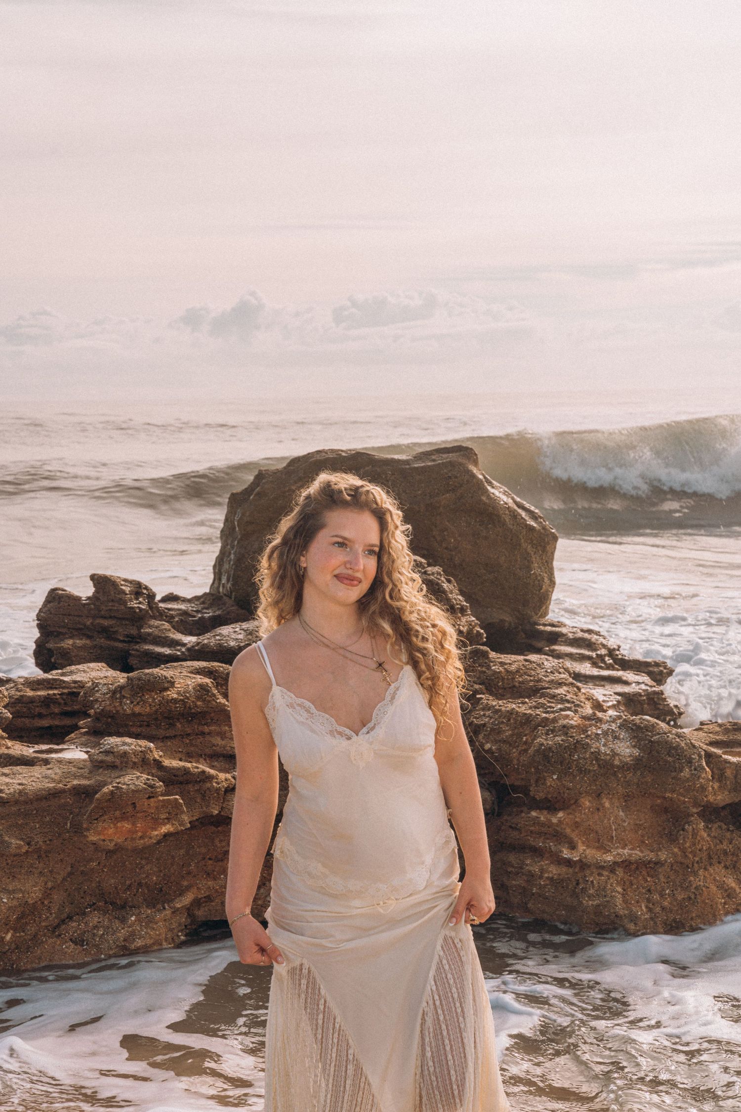 A woman in a white lace dress stands smiling on a rocky beach with ocean waves behind her.
