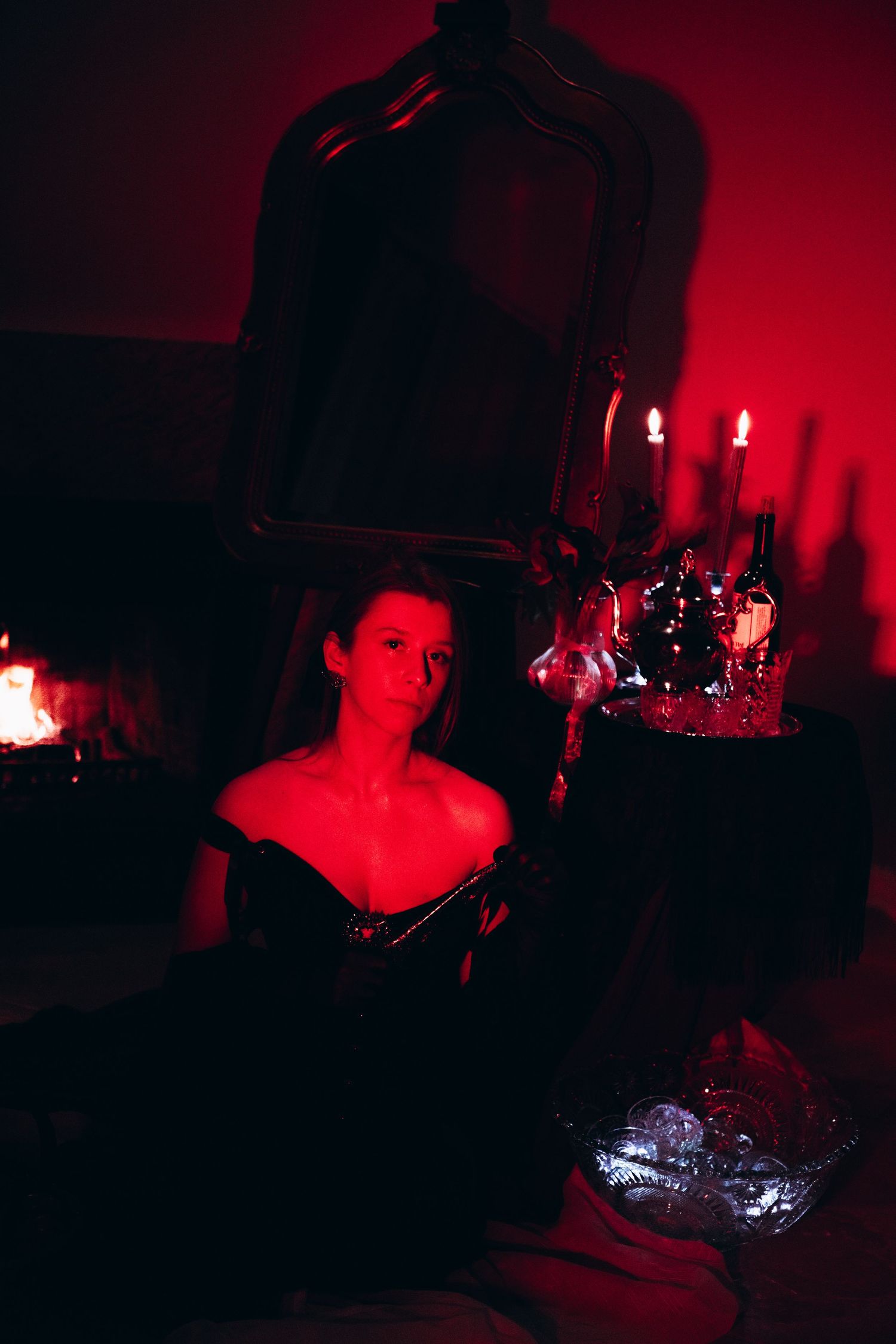 Woman in black off-shoulder dress posed by candlelit mirror in dramatic red-lit gothic setting.