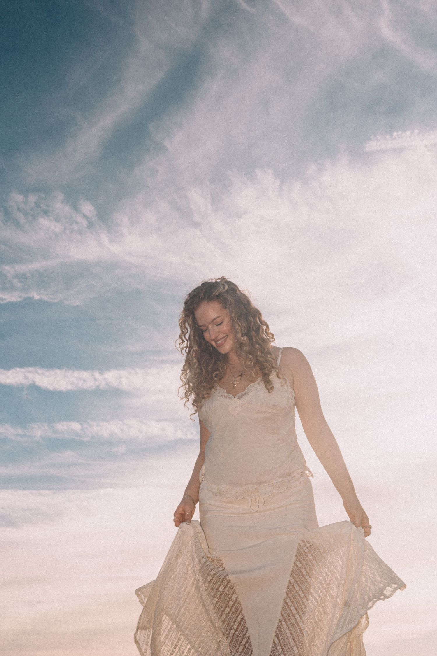 Woman in white lace dress holding skirt, curly hair, smiling outdoors against blue sky with wispy clouds.