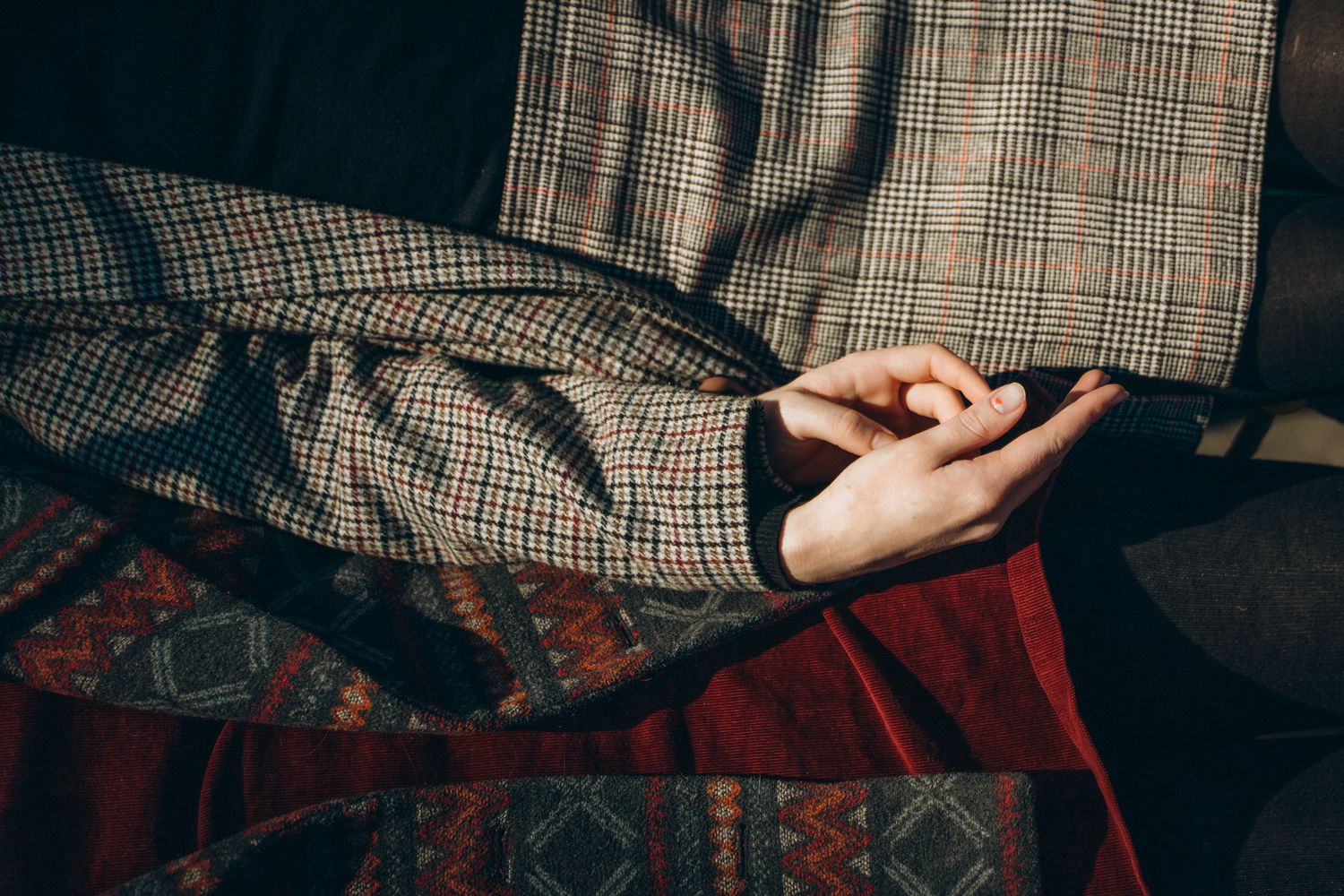 Clasped hands resting on plaid trousers with a colorful patterned textile in warm sunlight.