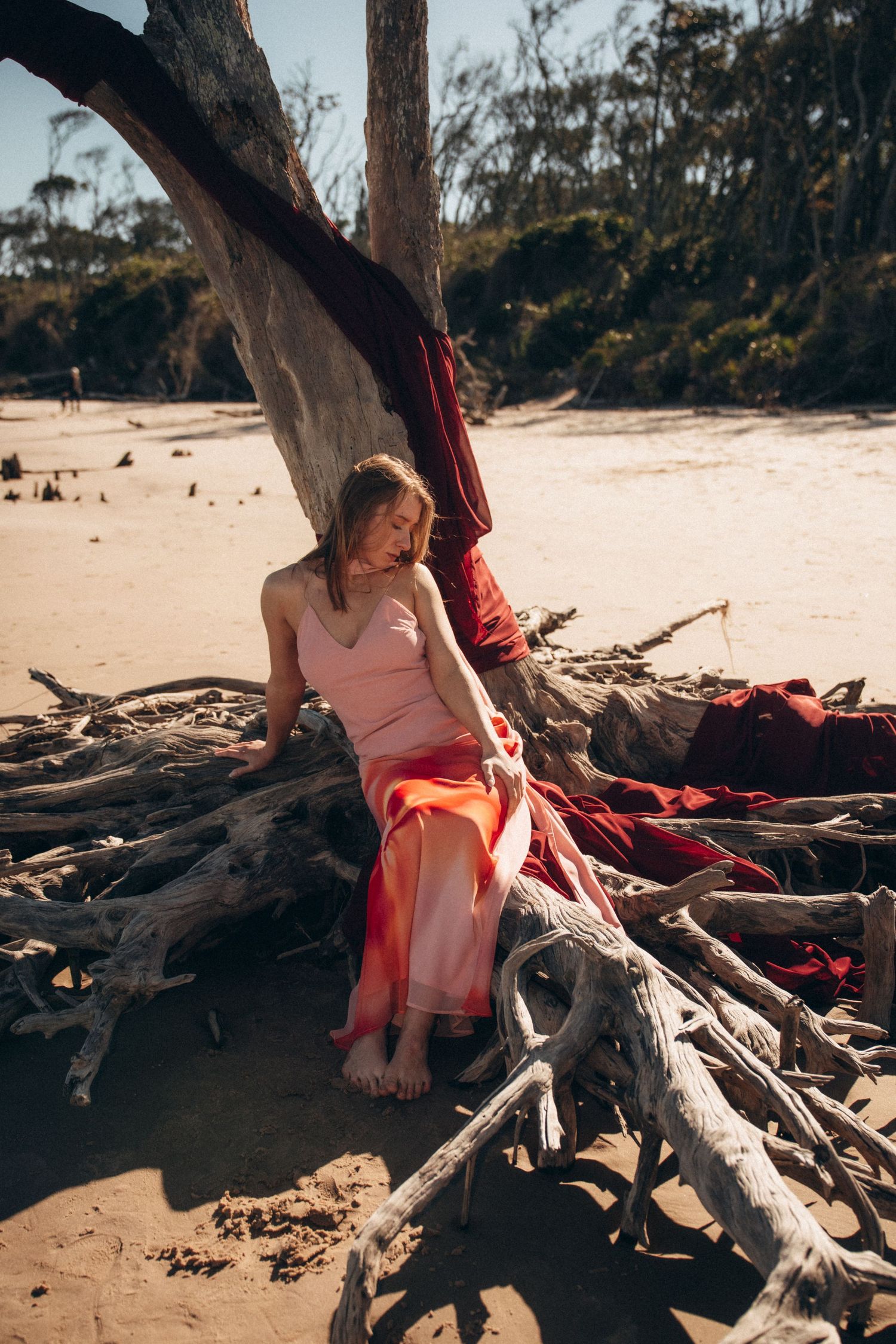 Someone in a pink dress sits on driftwood at a sandy beach with trees in the background.