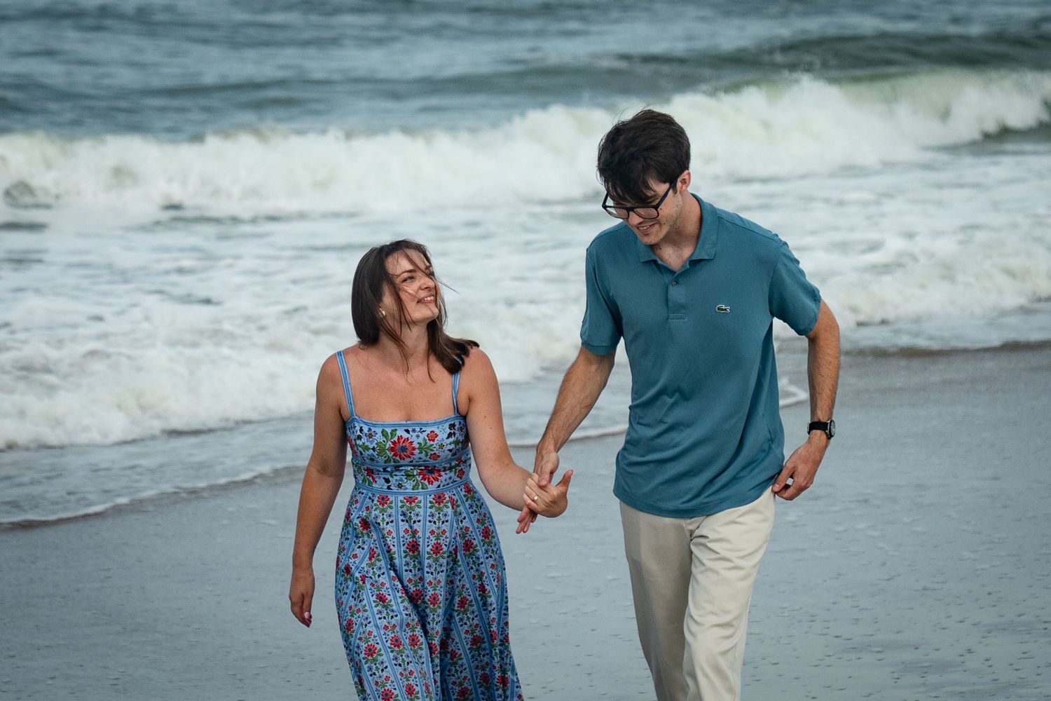 Couple walking together along a beach shoreline with waves crashing behind them at sunset.