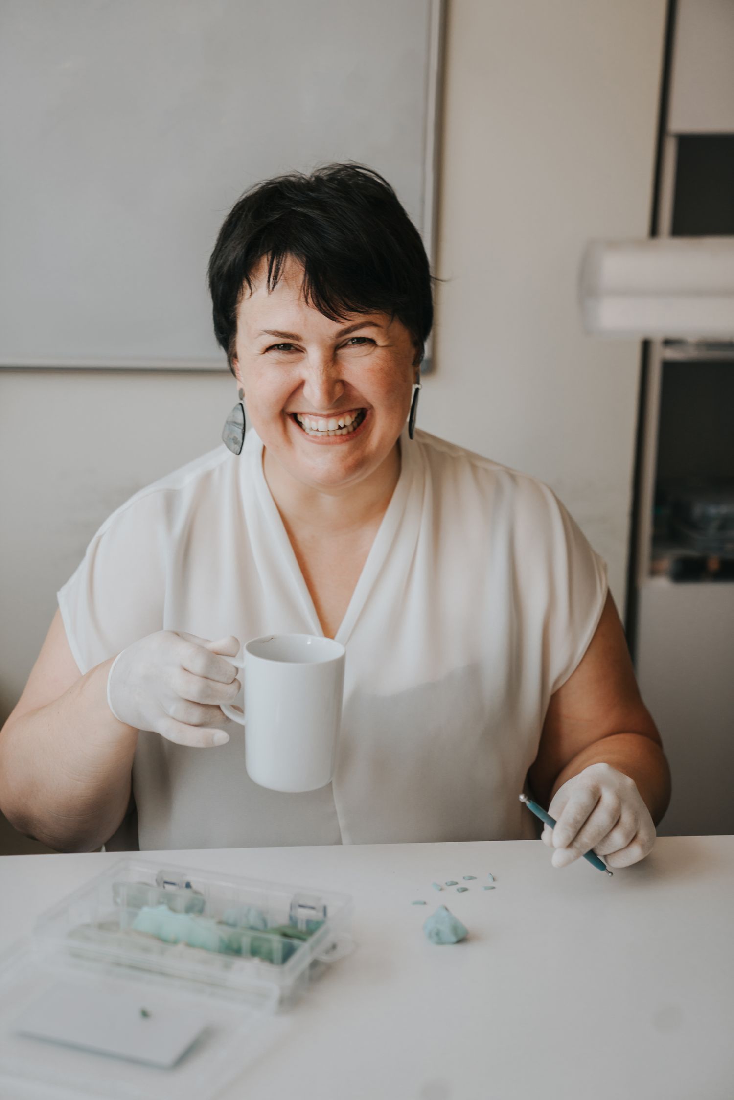 Smiling woman in white top and gloves holding a mug at a craft table with resin supplies.