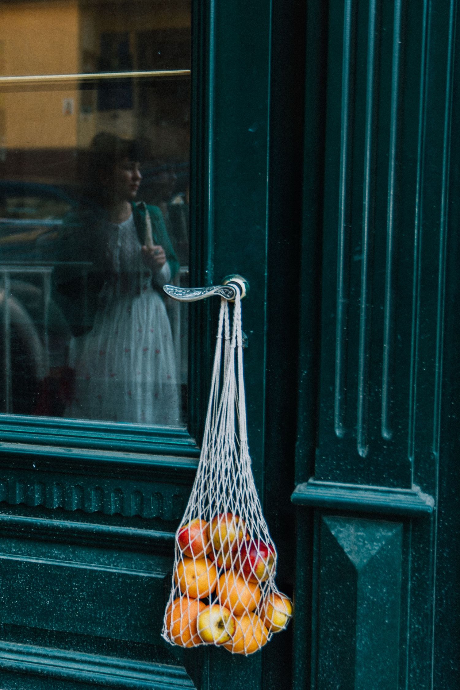 A white mesh string bag filled with fresh apples and oranges hanging on a dark green door with a woman reflected in the glass.