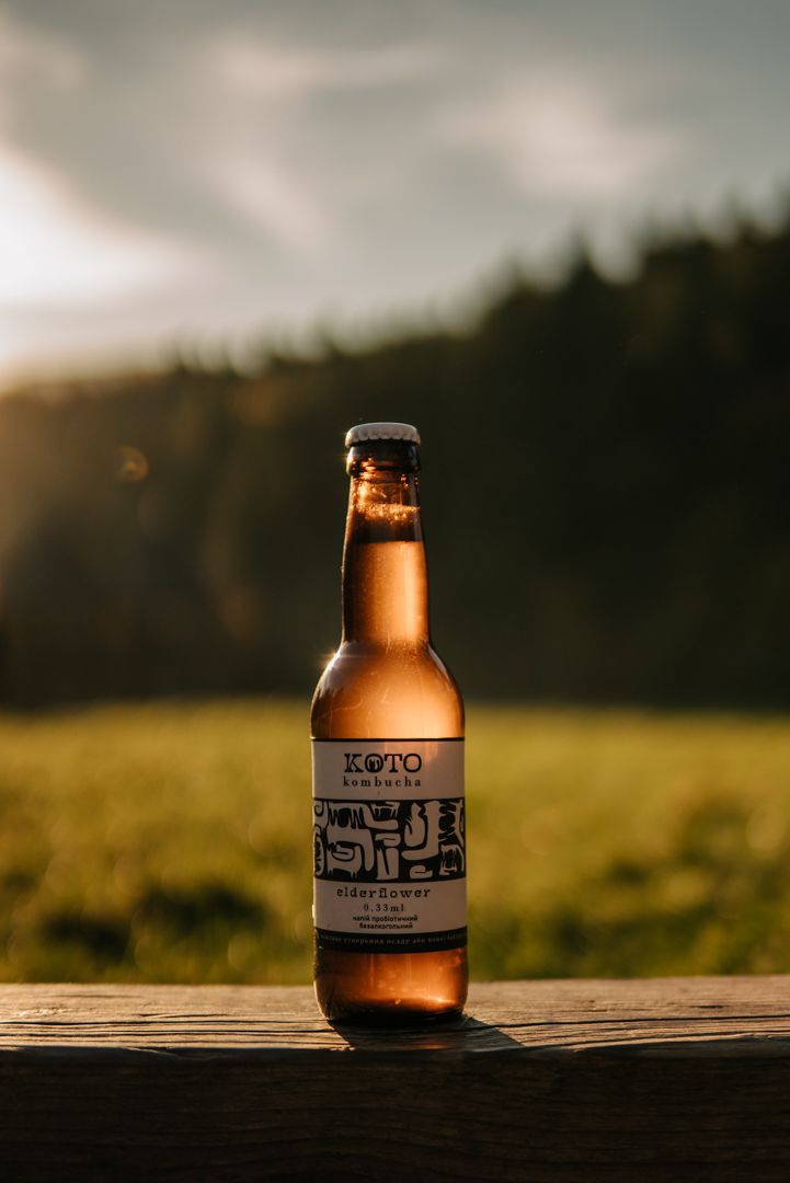 KOTO Kombucha elderflower bottle on wooden surface at golden hour with green meadow and forest backdrop.
