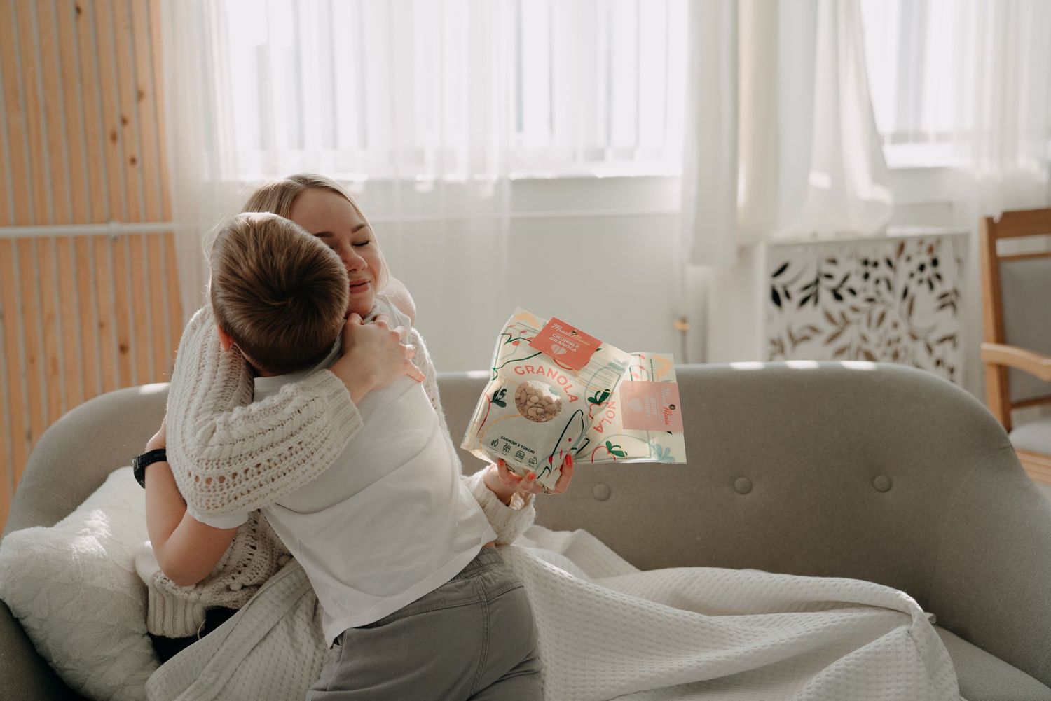 A mother hugging her child on a cozy sofa while holding a colorful gift box in a bright living room.