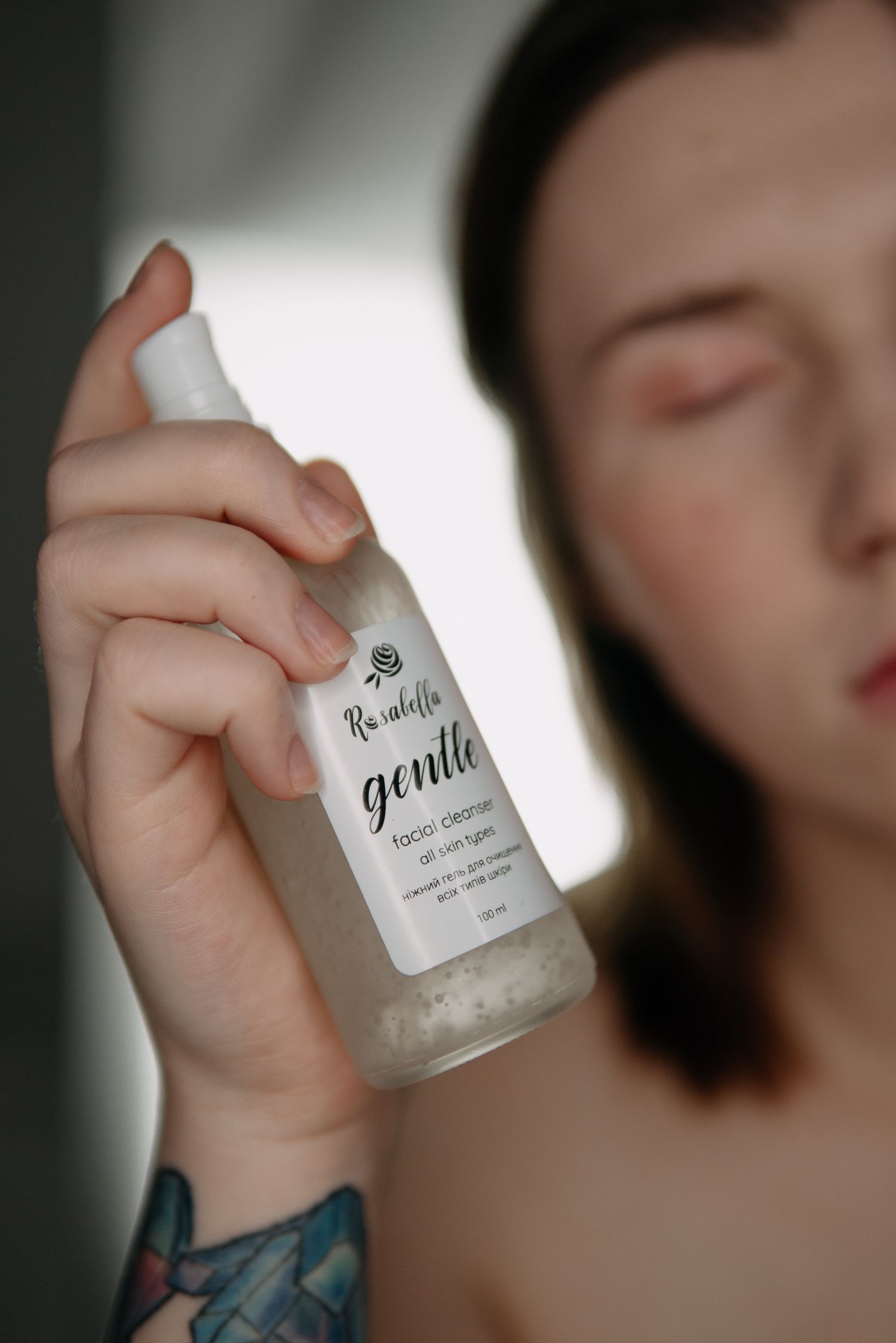 Woman with tattoo holding Rosabella gentle facial cleanser bottle, suitable for all skin types.