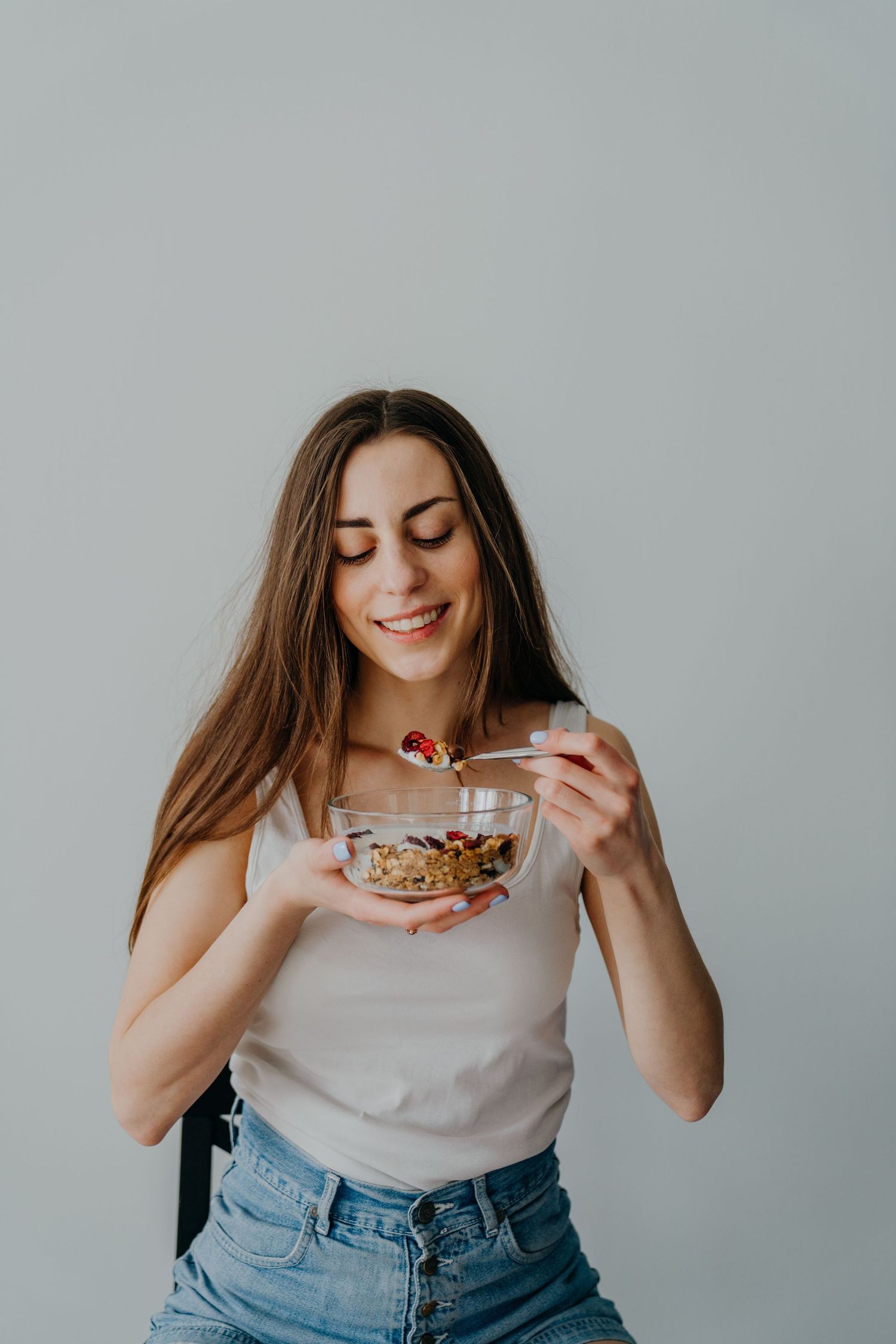 Smiling young woman with long hair eating granola from a glass bowl, wearing a white tank top and jeans.