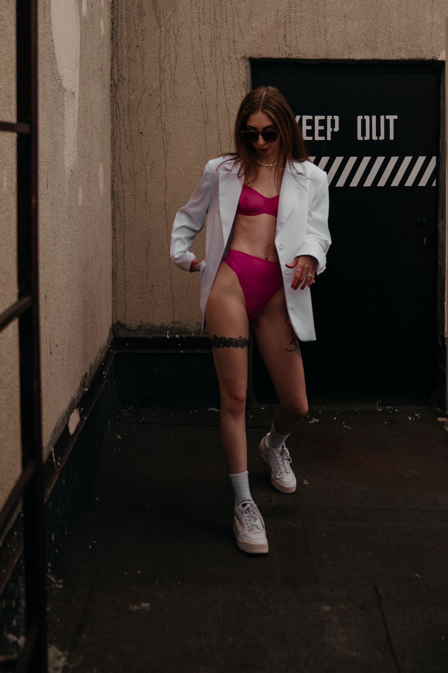 Woman in pink bikini and white blazer poses in urban alleyway wearing sneakers and sunglasses.