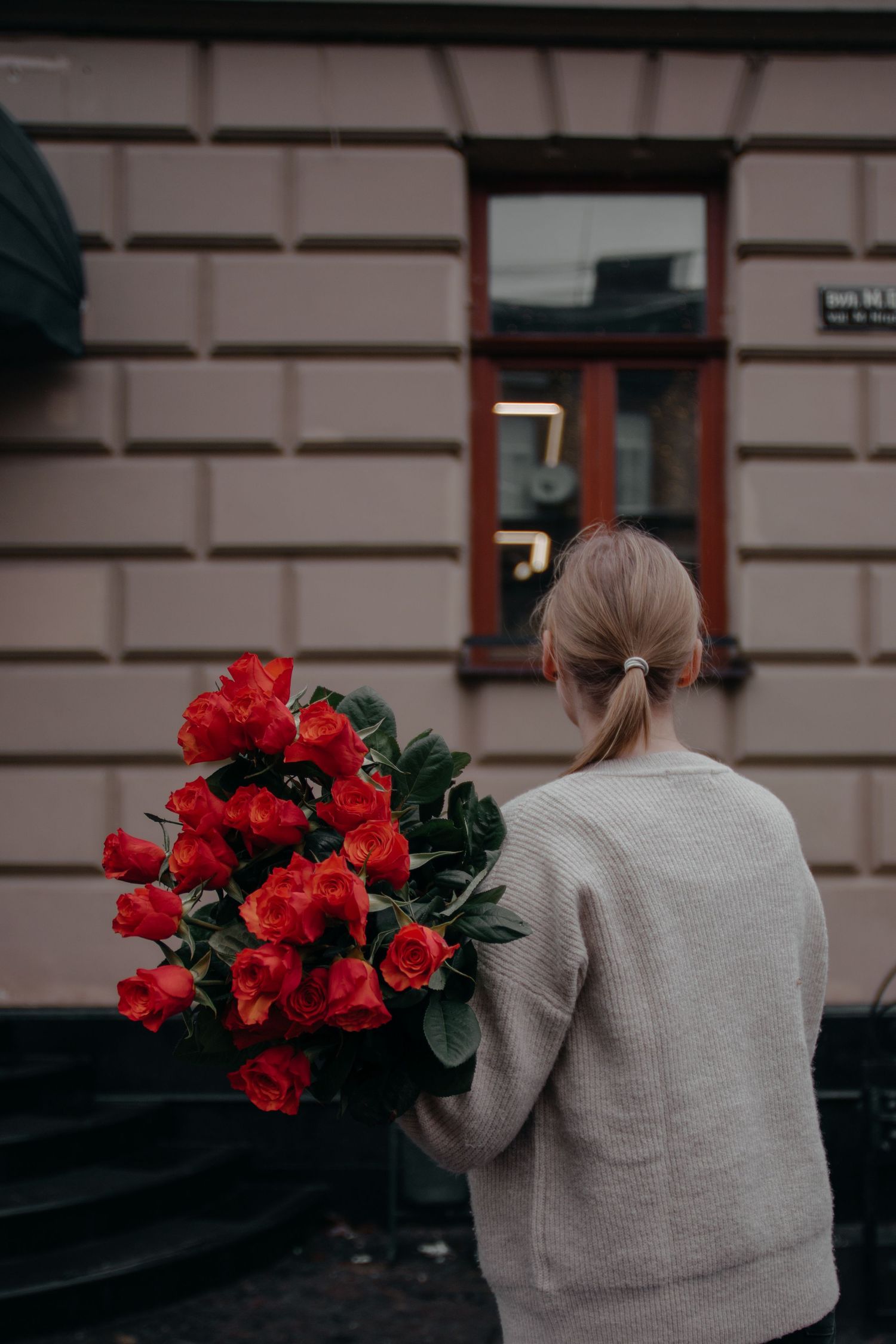 Woman with blonde hair in ponytail holding large bouquet of red roses, standing outside a building.