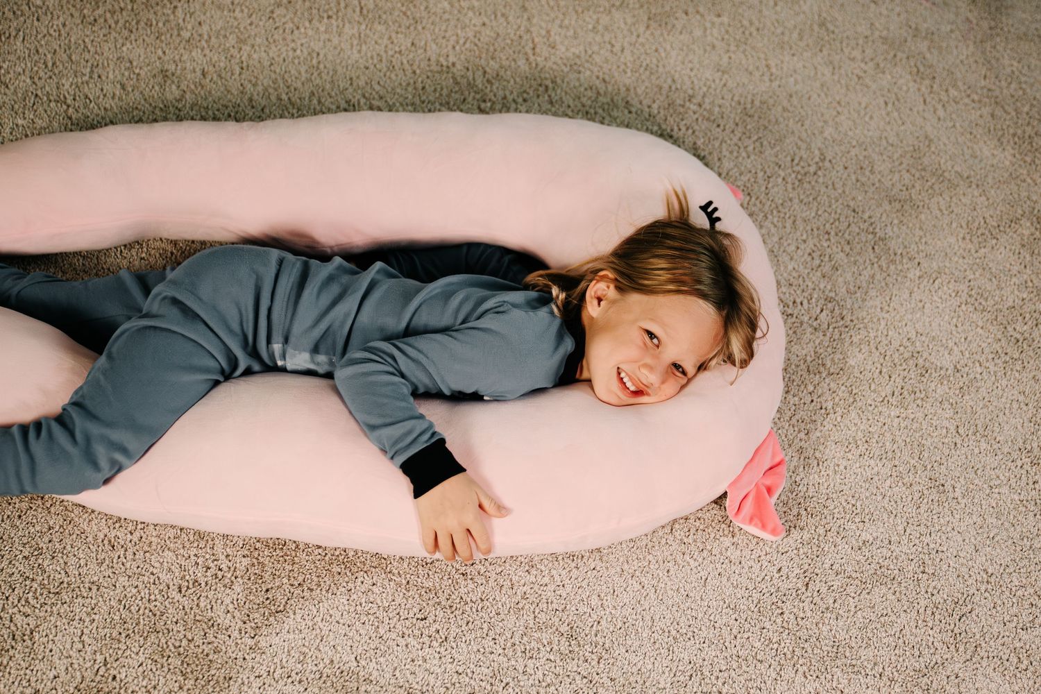 Young girl lying on a large pink U-shaped body pillow on a beige carpet, smiling up at the camera.
