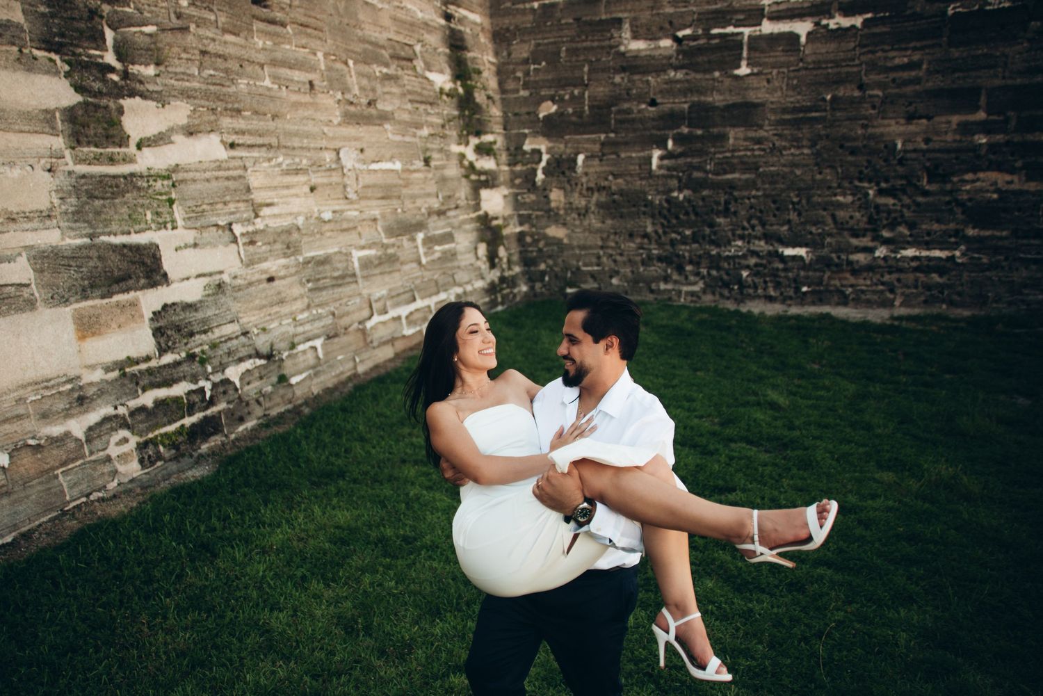 A groom carries his bride in white dress outdoors on green grass beside a stone brick wall.