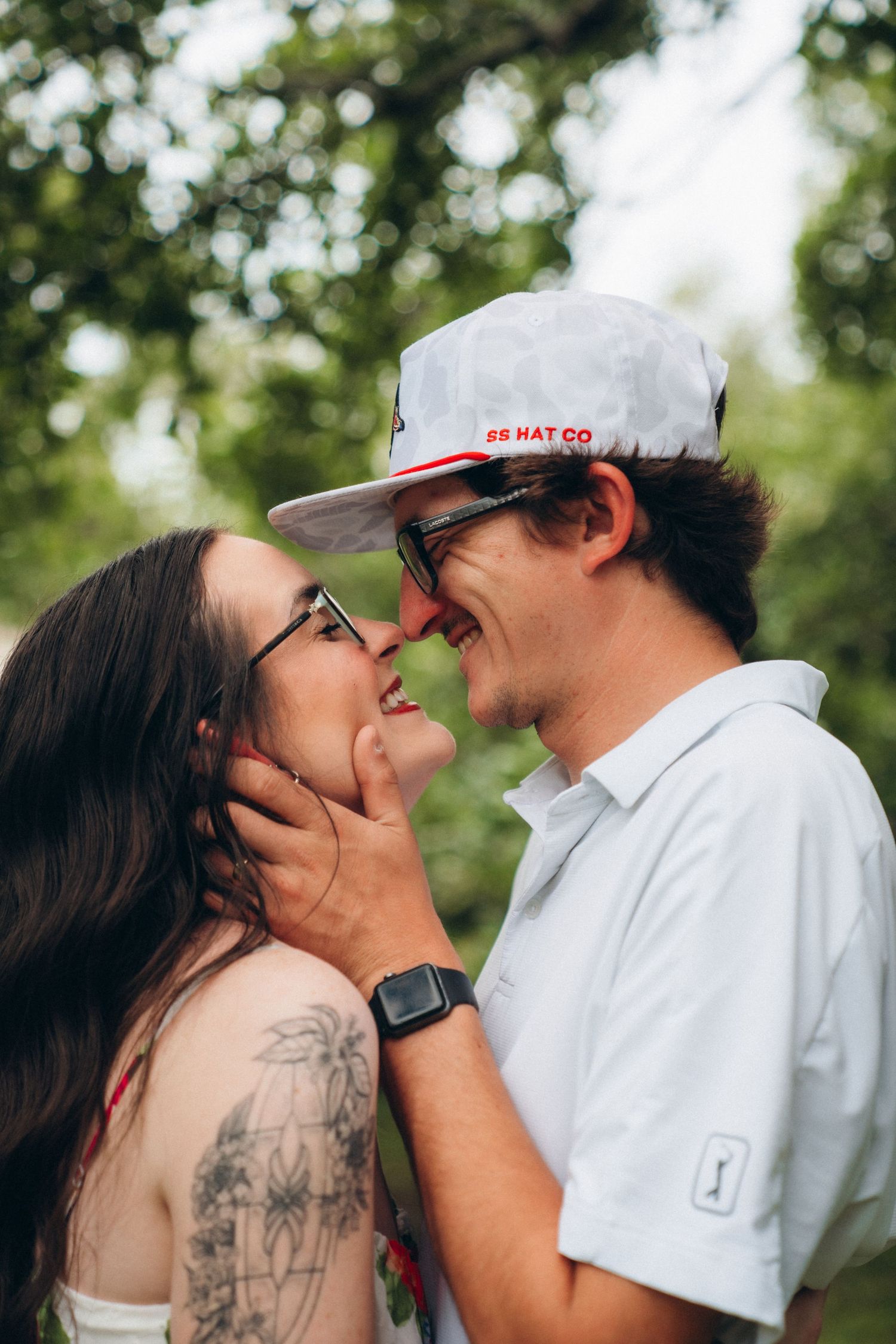 Smiling couple about to kiss outdoors, woman with long dark hair and tattoos, man wearing SS Hat Co white cap.