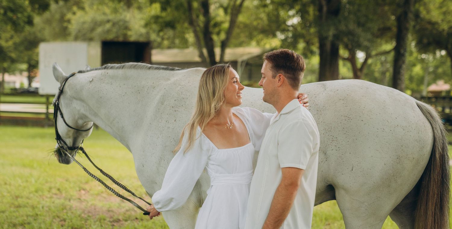 Couple in white outfits standing with a majestic white horse in a lush green field.