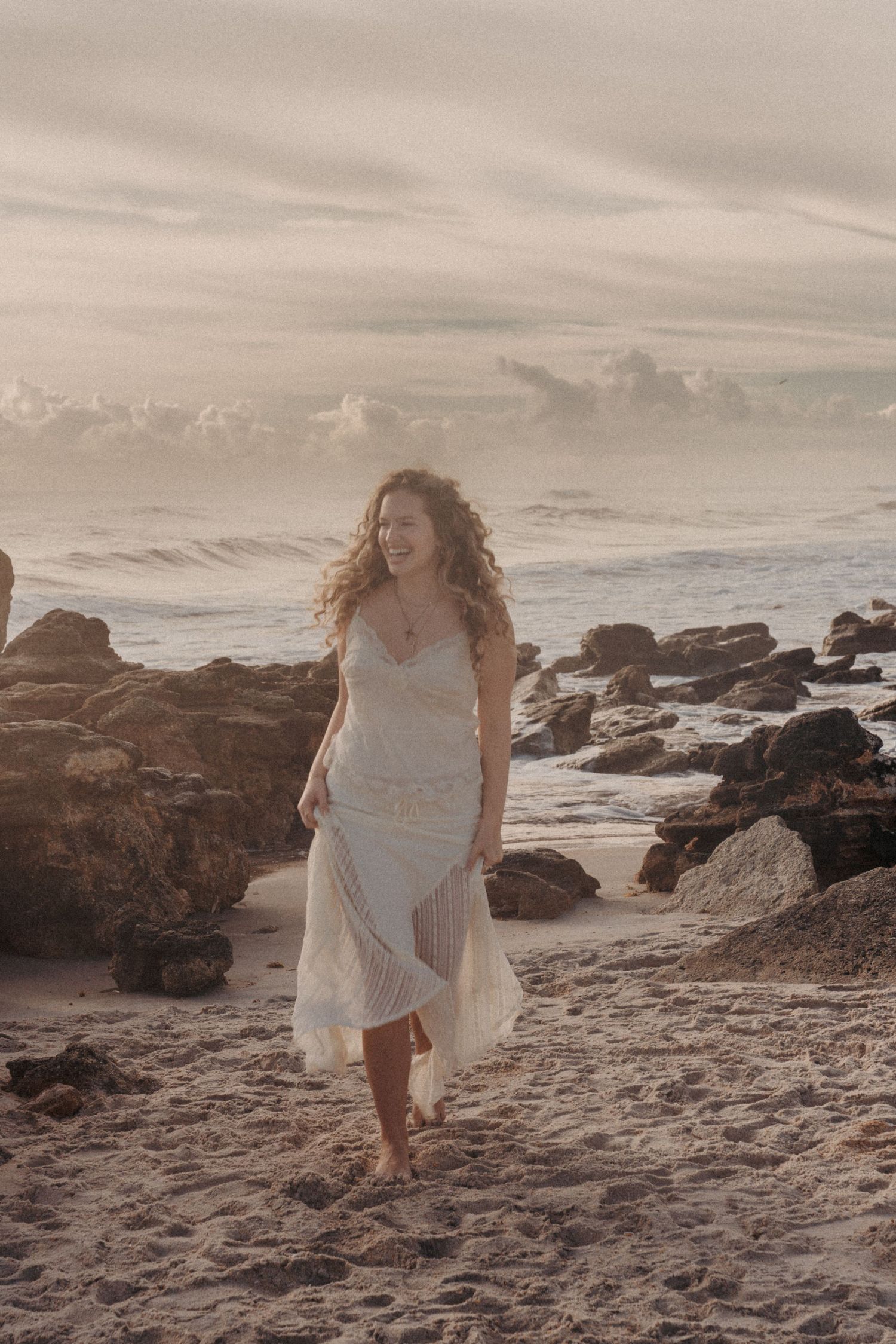 Woman in white dress walking barefoot on rocky beach at sunset with waves in background.