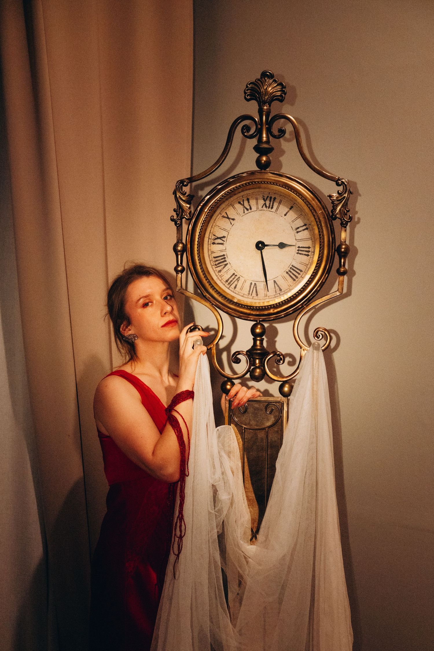 Woman in red dress standing beside ornate gold antique clock with white fabric draped over it.