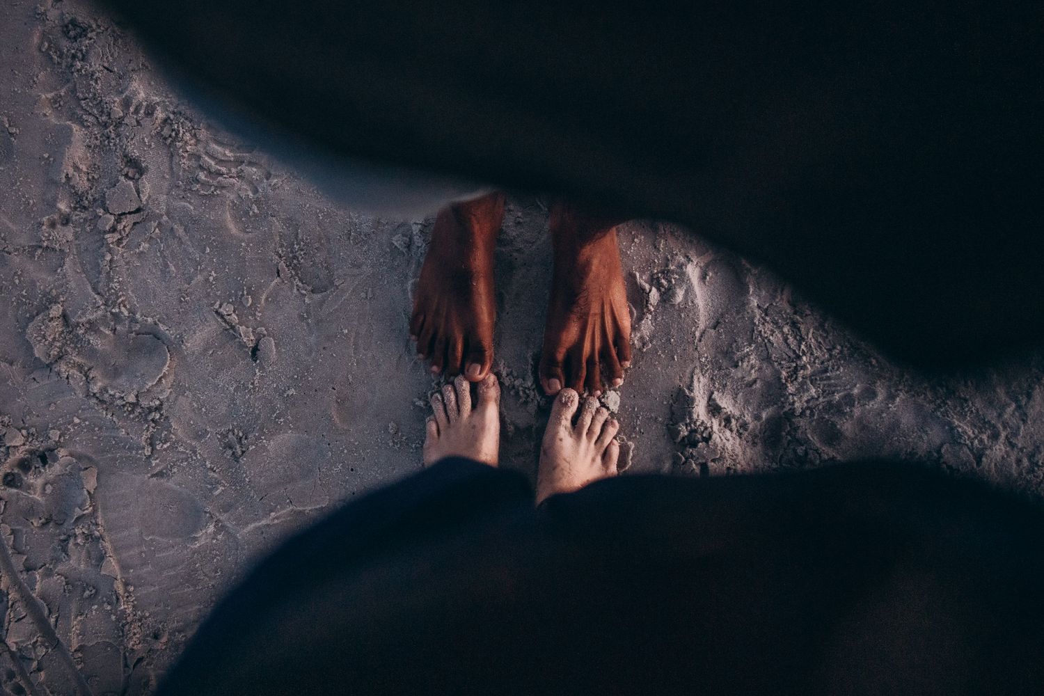 Two pairs of bare feet standing on wet sandy beach shore at dusk, captured from above.