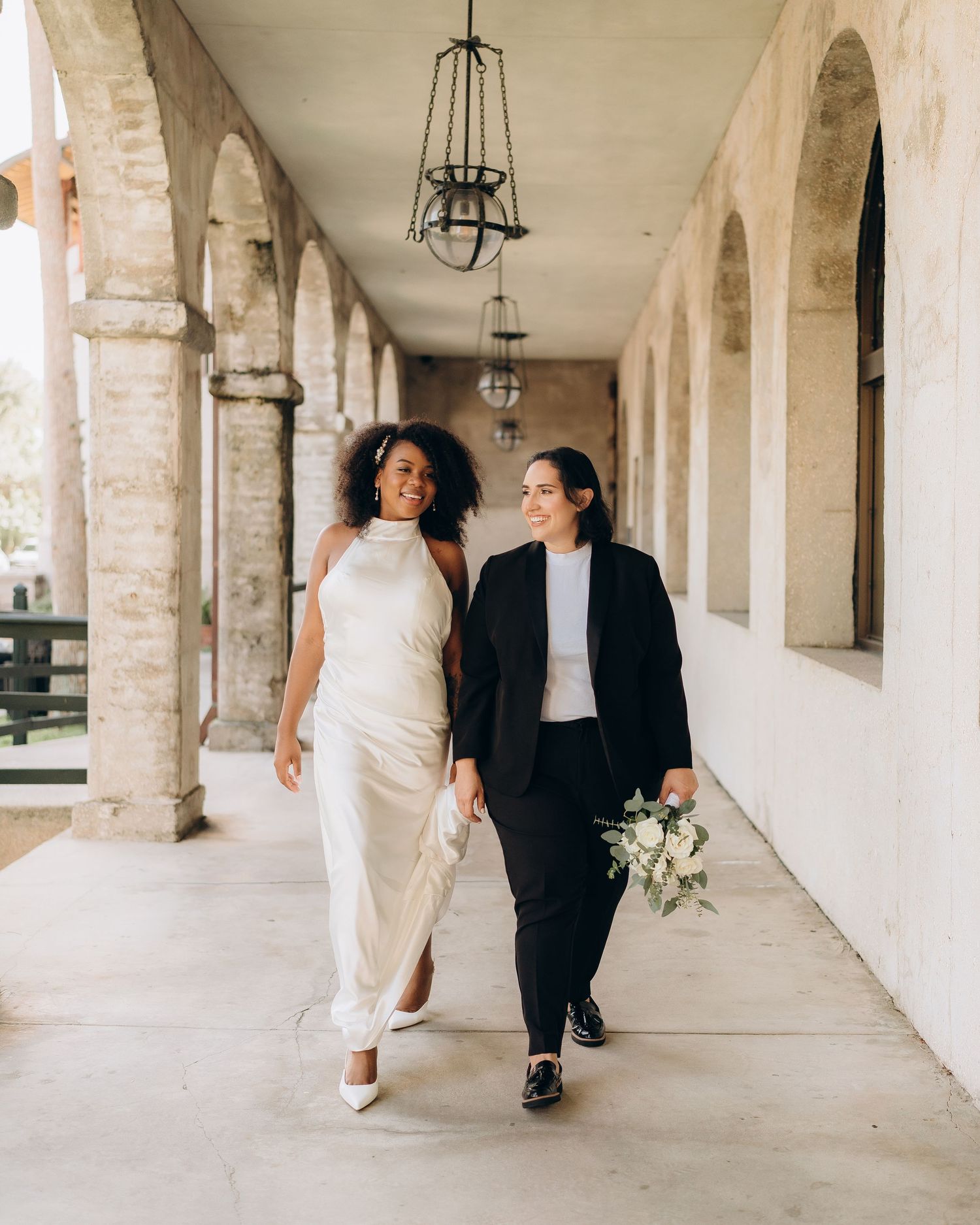Two brides walk together under arched colonnade, one in white gown and one in black suit holding bouquet.