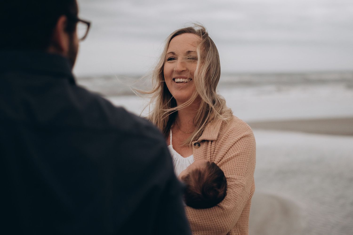 A smiling woman holds a newborn baby on a beach, laughing joyfully with a man in the foreground.