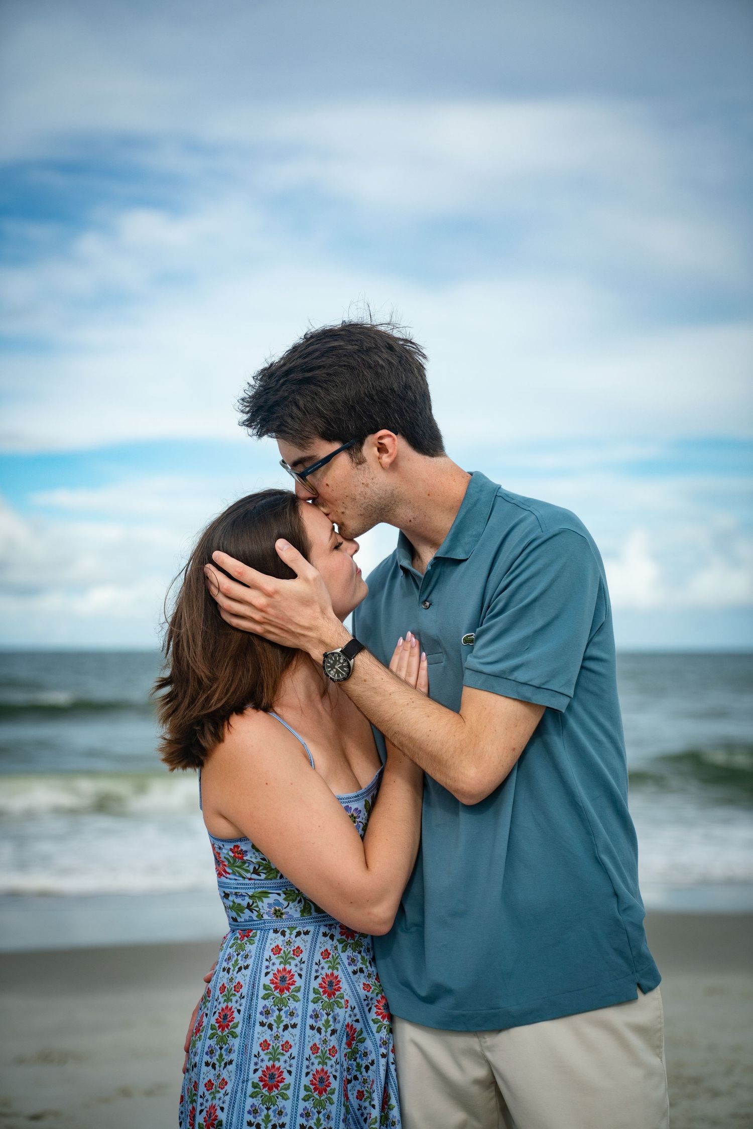 A romantic couple sharing a tender kiss on the beach with the ocean and cloudy blue sky in the background.