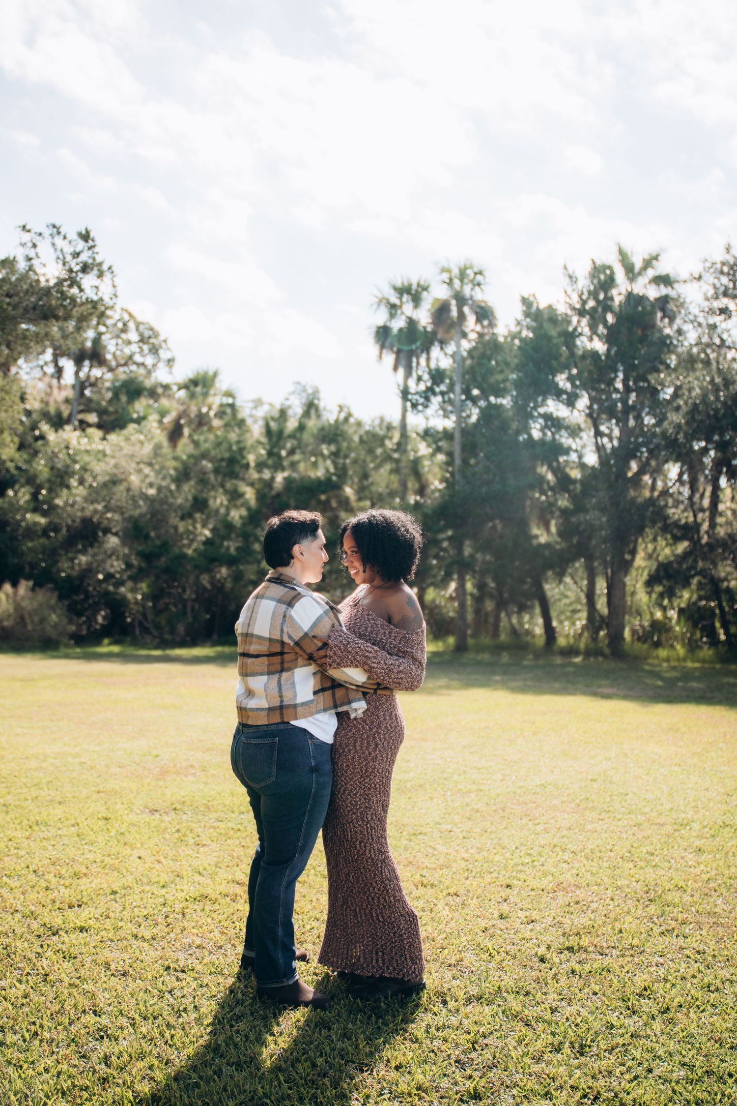 A couple shares a tender embrace while standing on a sunlit grassy field with palm trees in the background.
