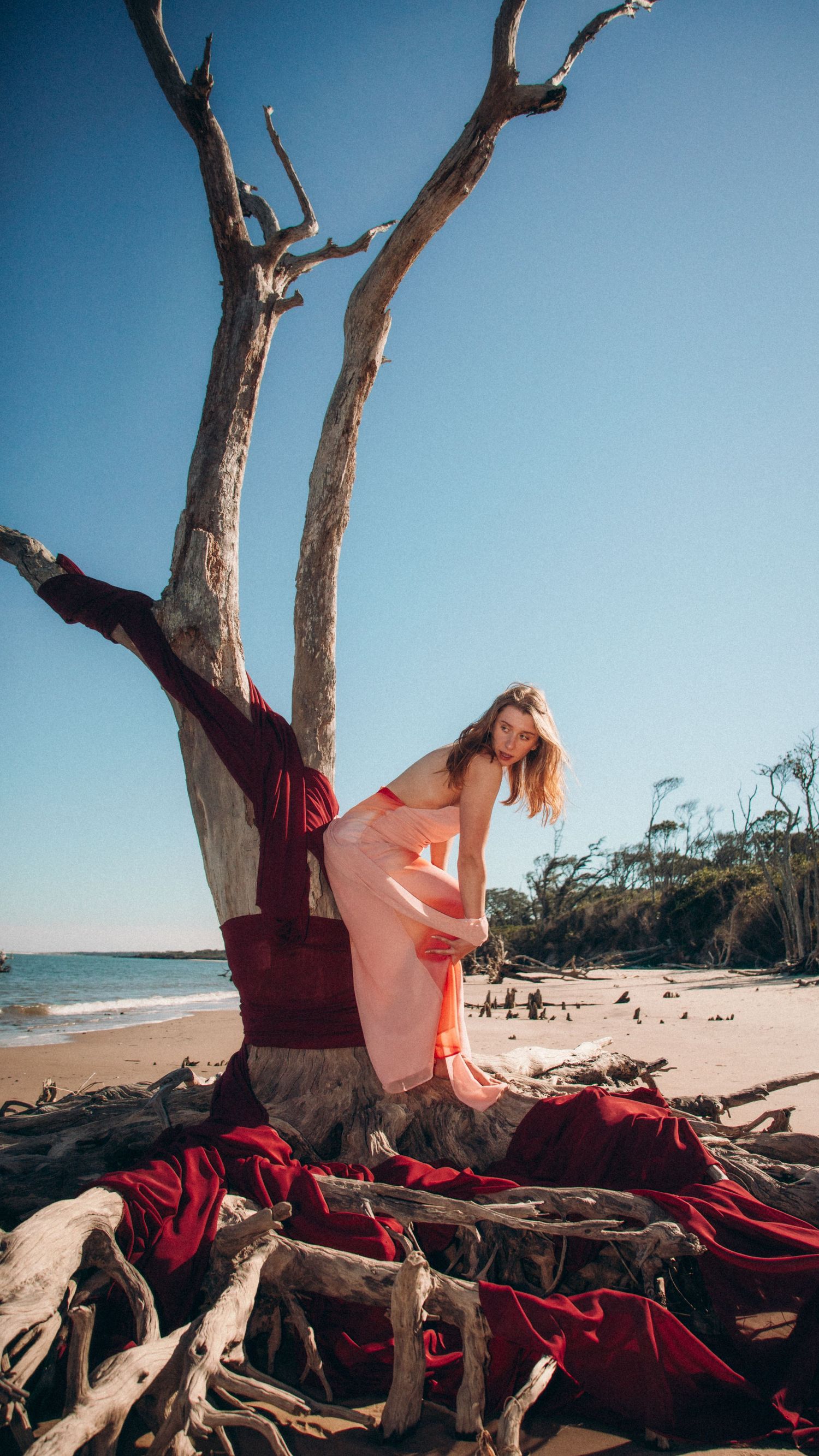 A coastal photography setup featuring red fabric draped over driftwood with a pink dress against the ocean backdrop.